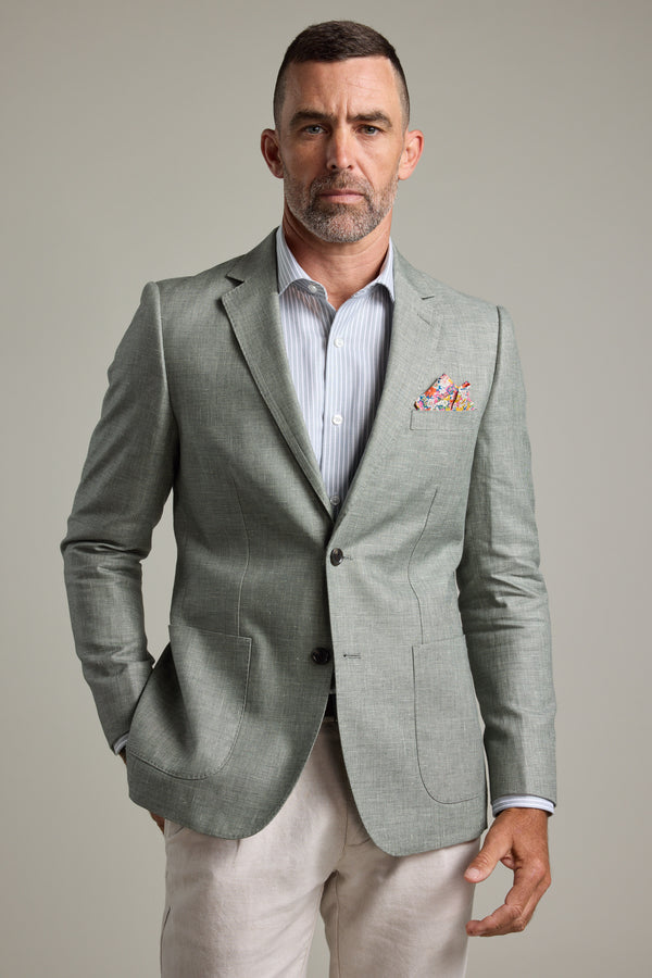 A man with short hair and a beard wears the Marion Texture Blazer by Barkers in light gray over a striped shirt, with a colorful pocket square and light trousers—an ideal summer occasion look, standing against a plain background.