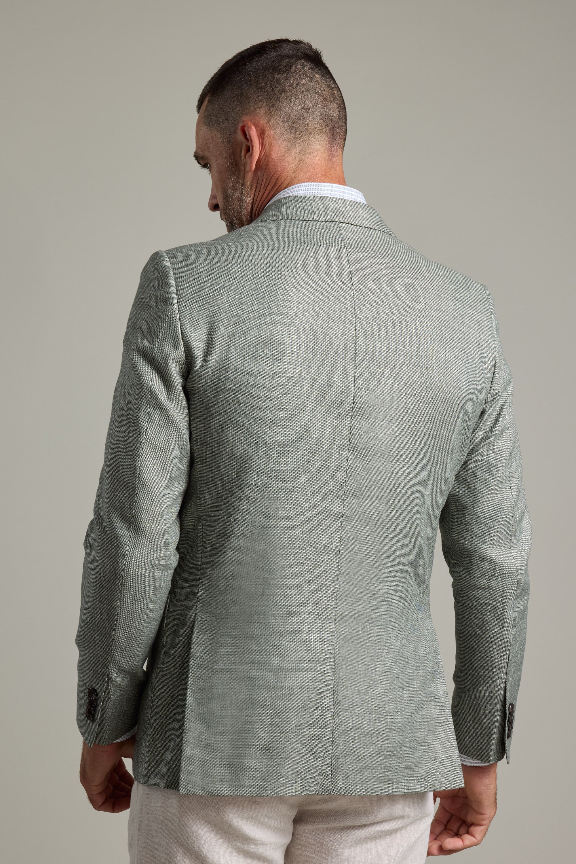 A man with short hair and a trimmed beard is seen from the back, wearing the Barkers Marion Texture Blazer in light gray and white pants, standing against a plain light gray background.