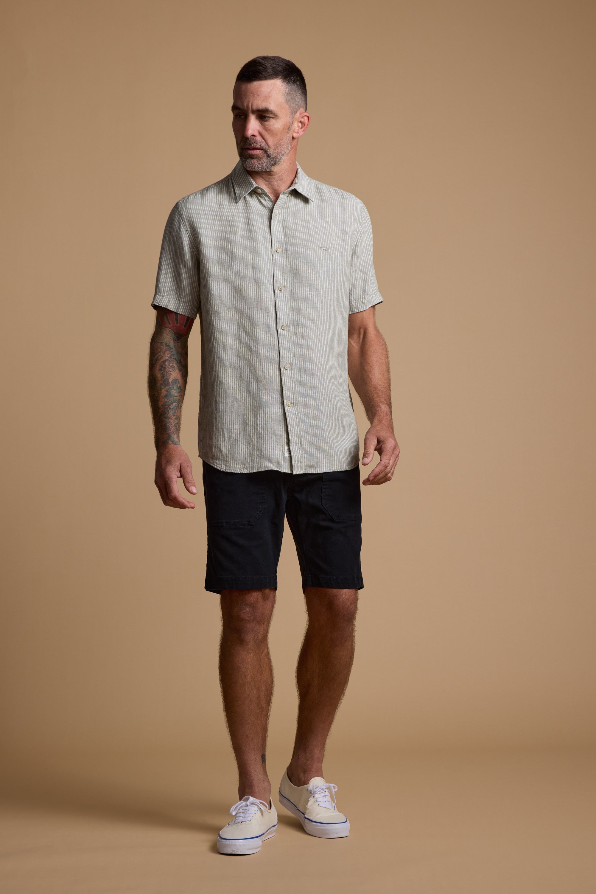A man stands against a beige background wearing Barkers Tidal GD Utility Shorts, a light cotton short-sleeve button-up shirt, and white sneakers. He has tattoos on his left arm and gazes slightly to the side.