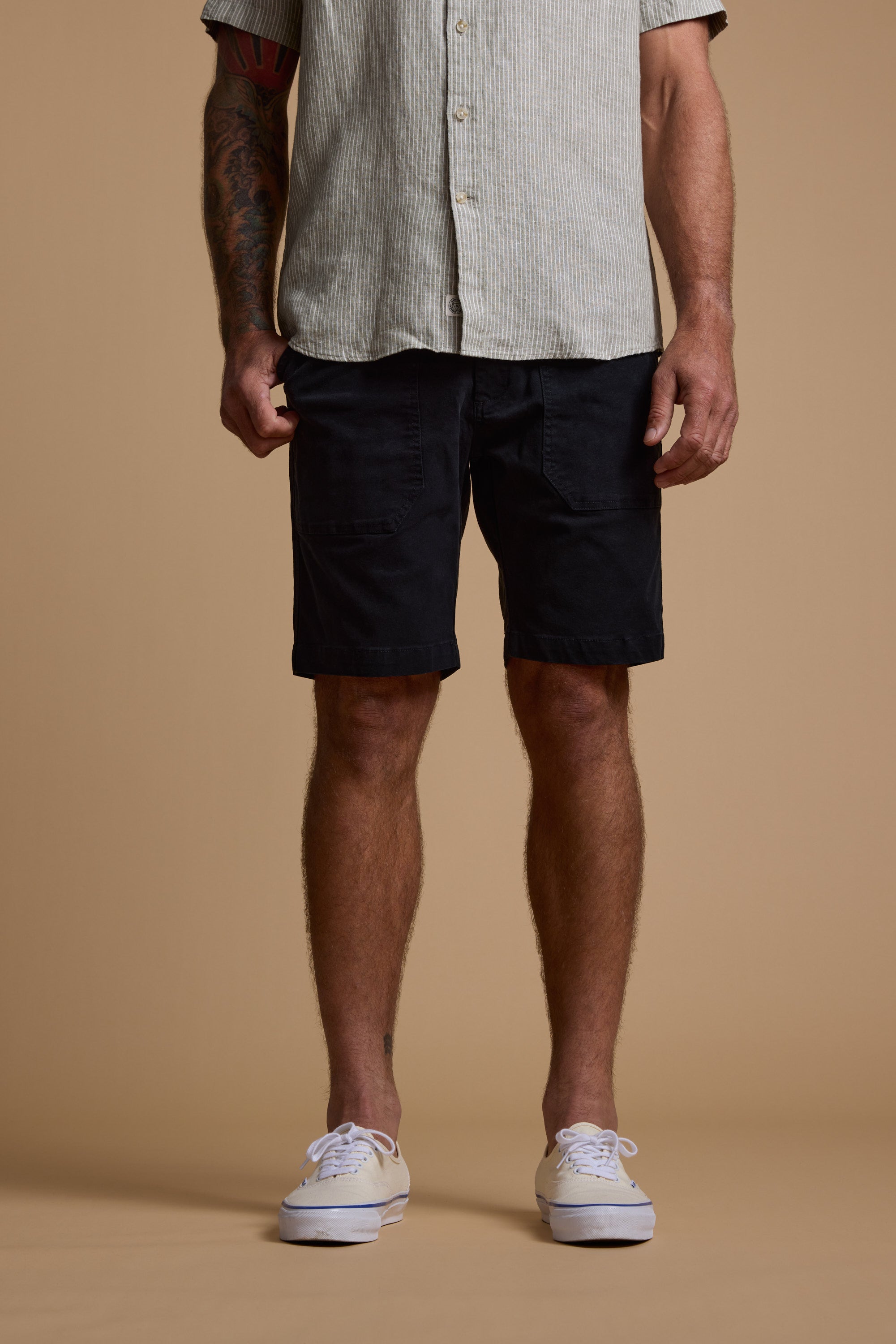 A man stands against a plain beige background, shown from shoulders to feet, wearing a modern fit light striped button-up shirt, Barkers Tidal GD Utility Shorts in black, and white sneakers.