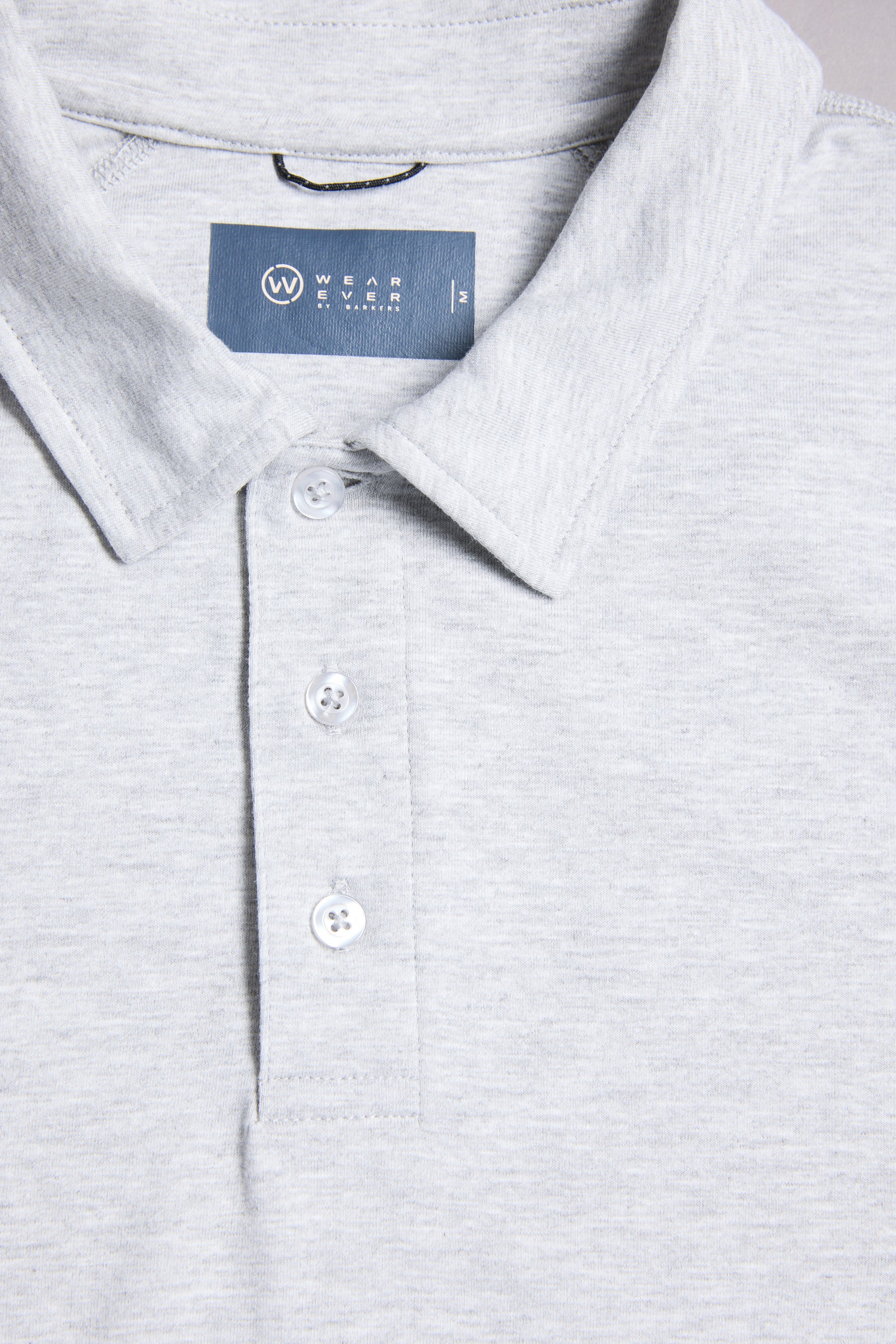 Close-up of the WearEver Outbound Polo in light grey, made from breathable cotton-blend fabric. Features a collar, three white buttons, and a blue inner label with the WearEver brand name.