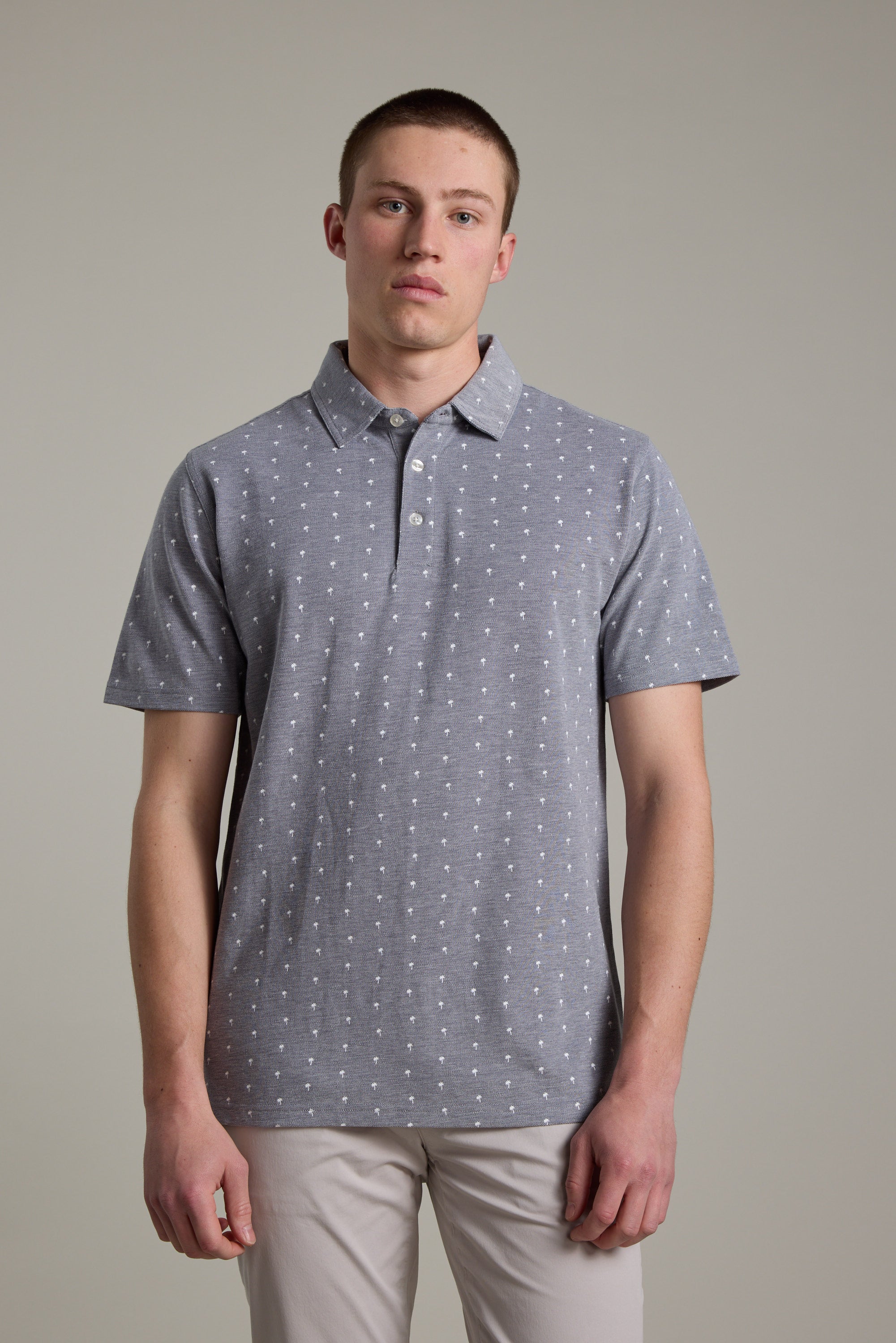 A young man faces forward in the Barkers Melrose Pique Polo—an organic cotton shirt with a subtle white dot pattern—paired with light pants, showcasing effortless summer style against a plain light gray background.