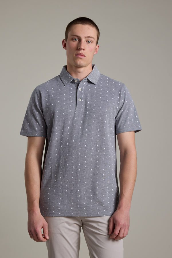 A young man faces forward in the Barkers Melrose Pique Polo—an organic cotton shirt with a subtle white dot pattern—paired with light pants, showcasing effortless summer style against a plain light gray background.