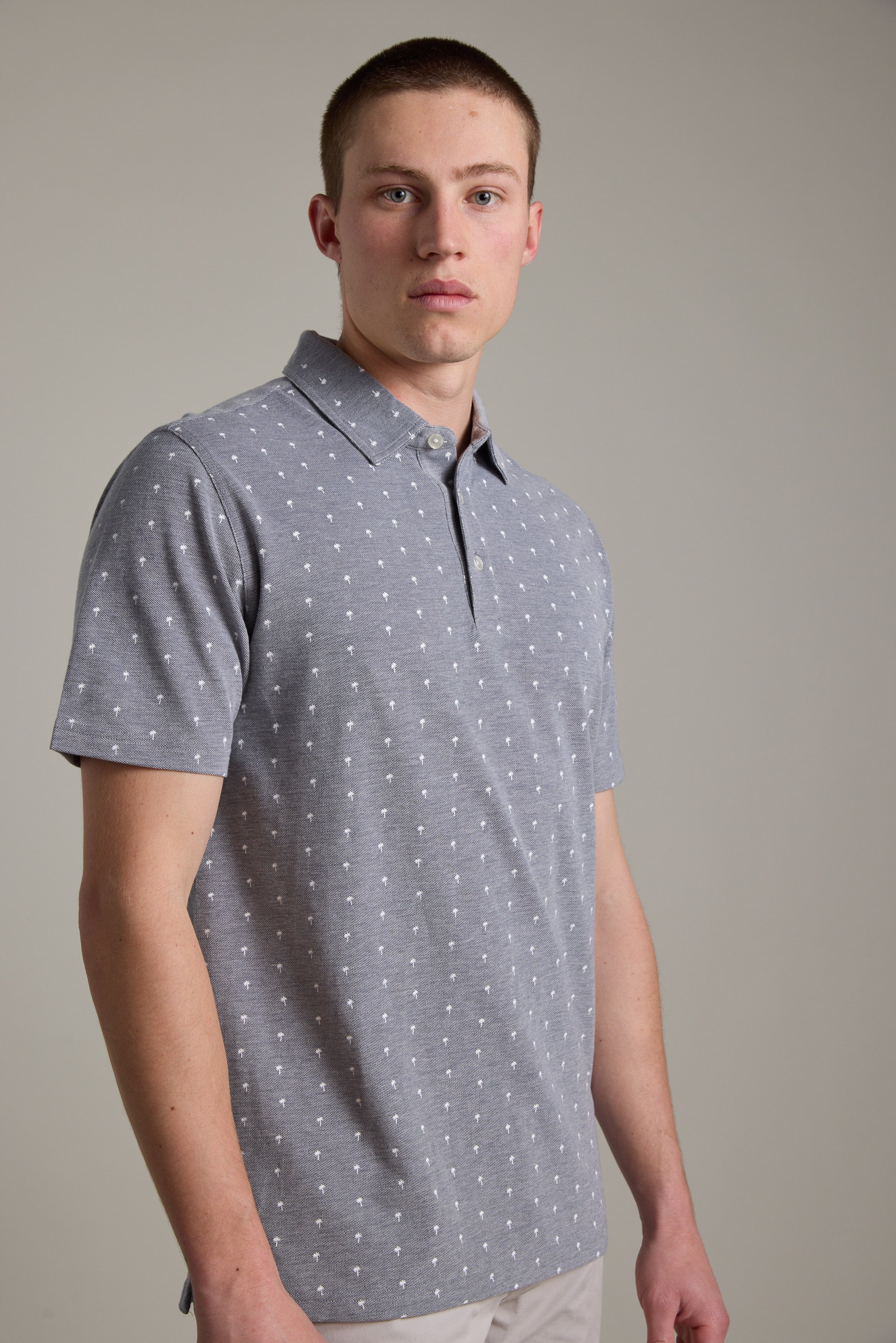 A young man with short hair models the Barkers Melrose Pique Polo in light gray with a subtle white dot pattern, set against a plain background—an ideal choice for effortless summer style.