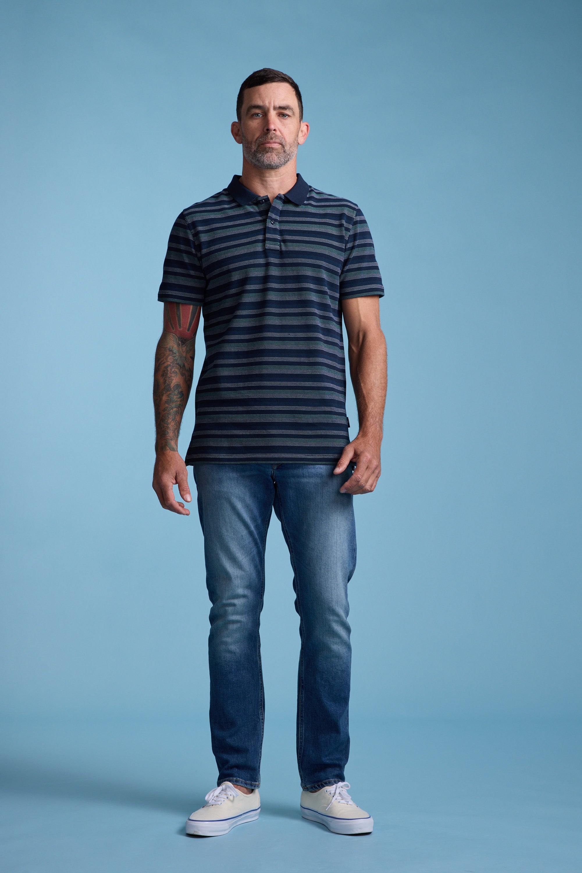 A man with short hair and a tattoo on his left arm stands facing forward, wearing the Barkers Dalton Stripe Polo with blue jeans and white sneakers against a plain light blue background.