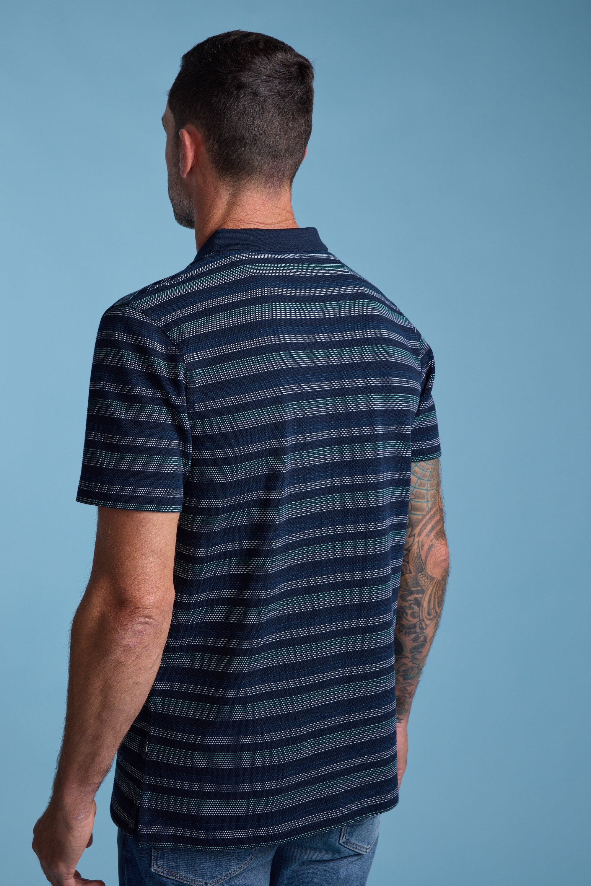 A man with short dark hair wears the Barkers Dalton Stripe Polo and jeans, standing with his back to the camera on a light blue background. His right arm shows off a colorful tattoo sleeve—great for easy summer style inspiration.