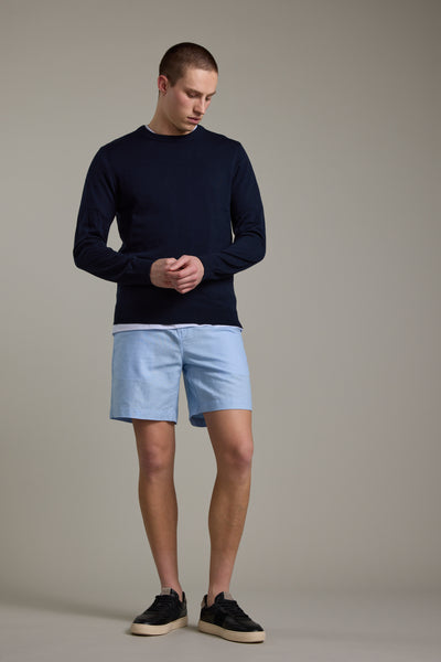 A man stands in a studio wearing a navy sweater, black sneakers, and Barkers Jasper Linen Weekend Short in light blue. He looks down with his hands clasped in front of him against a plain gray background.