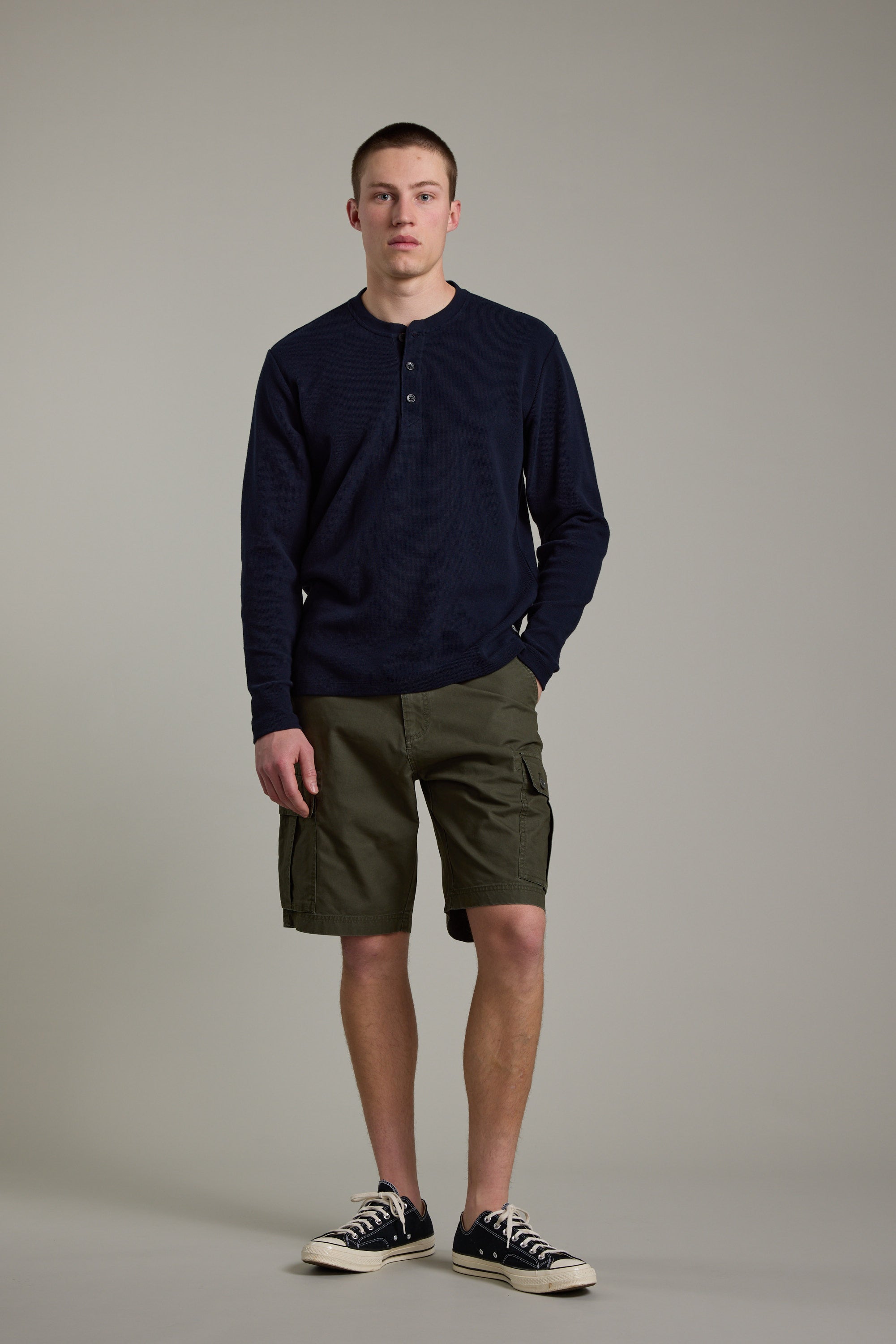 A young man stands against a plain background, wearing Barkers' Avenue Waffle Henley in dark navy, paired with olive cargo shorts and black-and-white sneakers. One hand is in his pocket as he looks straight ahead.
