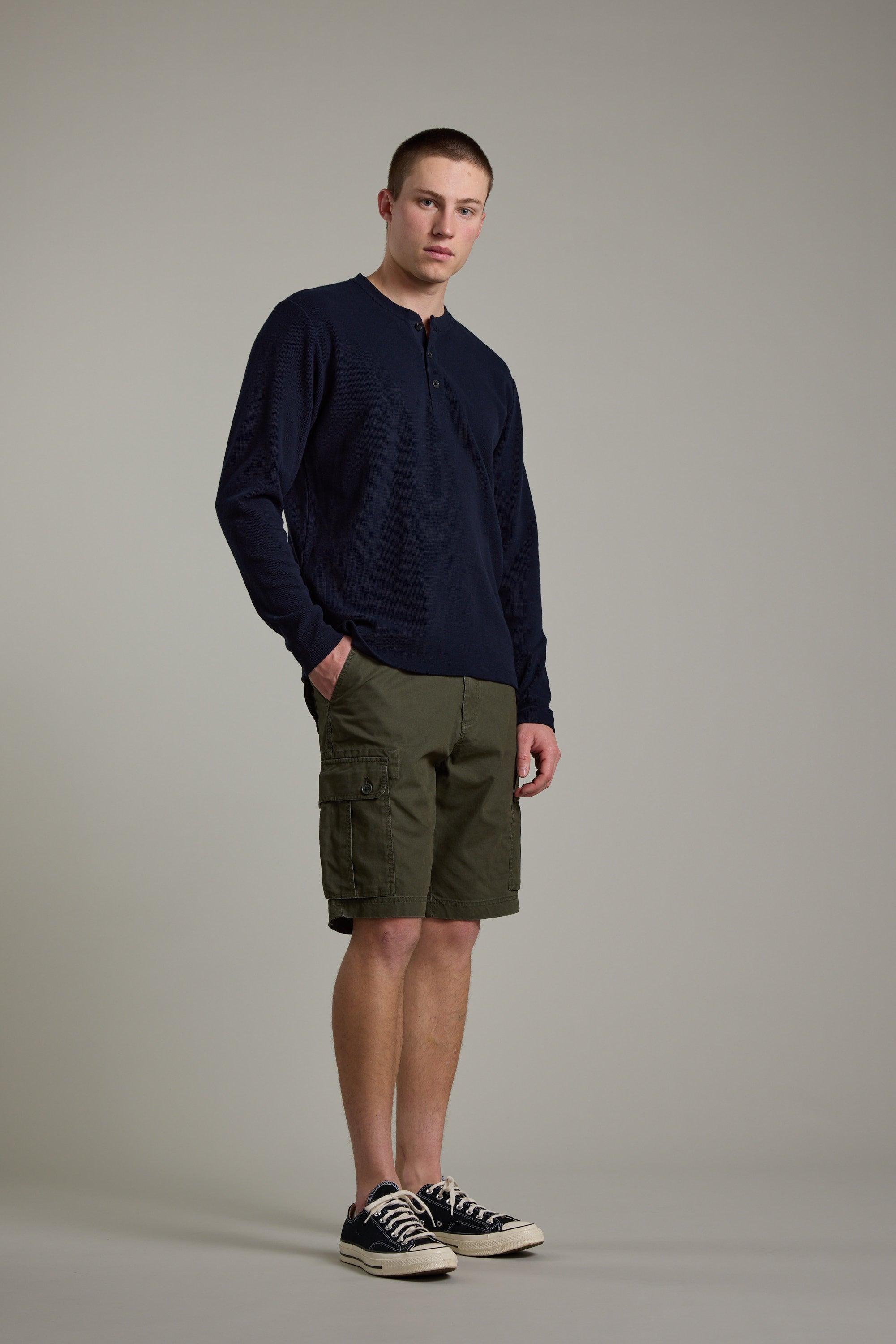 A young man with short hair and a neutral expression stands against a plain background, wearing the Barkers Avenue Waffle Henley in navy, olive cargo shorts, and black low-top sneakers, with his hands in his pockets.