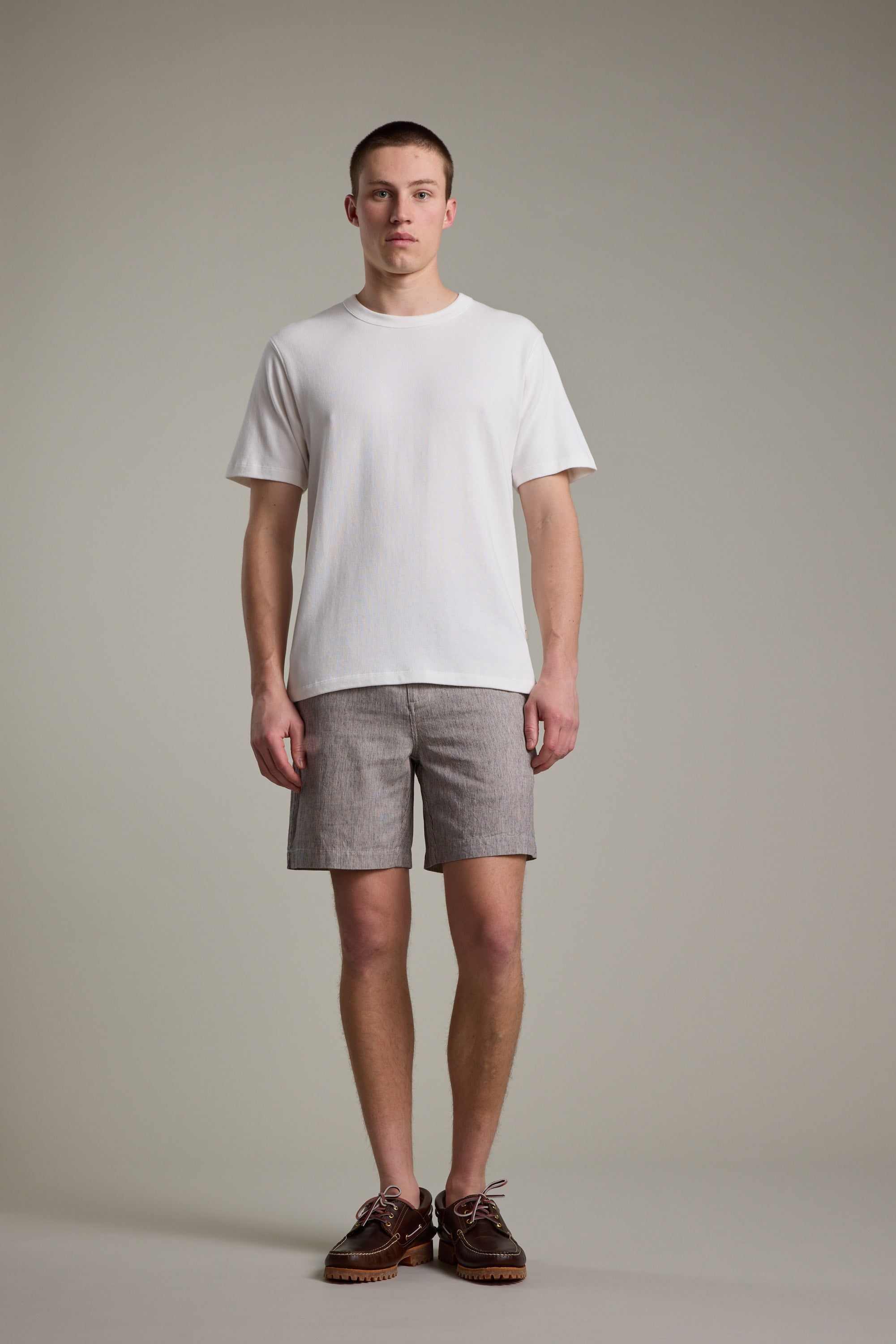 A young man stands facing forward against a light gray background, wearing Barkers' Woodley Weekend Short with an elastic waistband, a plain white T-shirt, and brown shoes. He has short hair and a neutral expression, arms relaxed at his sides.