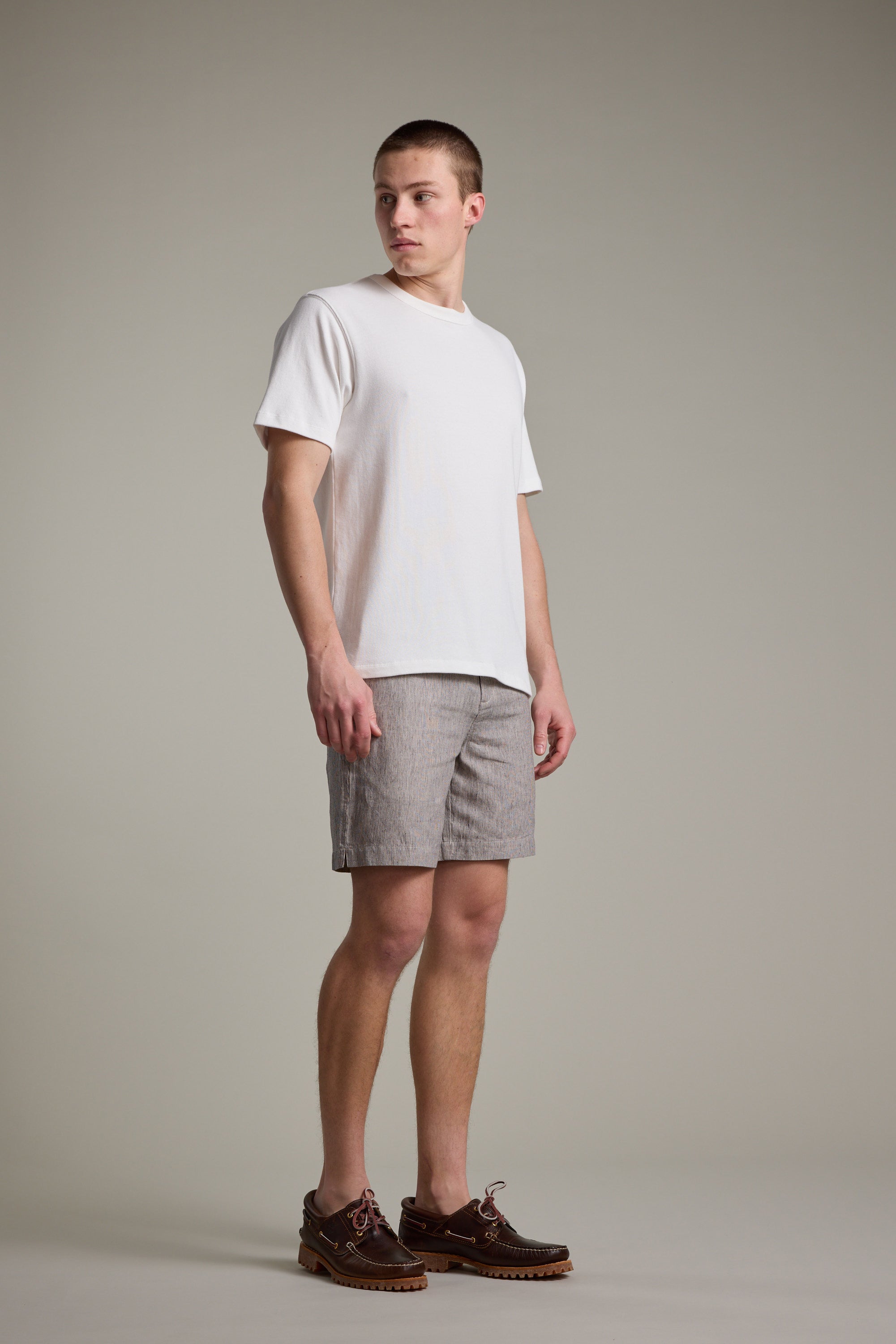 A young man stands against a plain background, wearing a white t-shirt and Barkers Woodley Weekend Shorts in light gray, paired with dark brown boat shoes featuring red laces. He looks slightly to his left with a neutral expression.