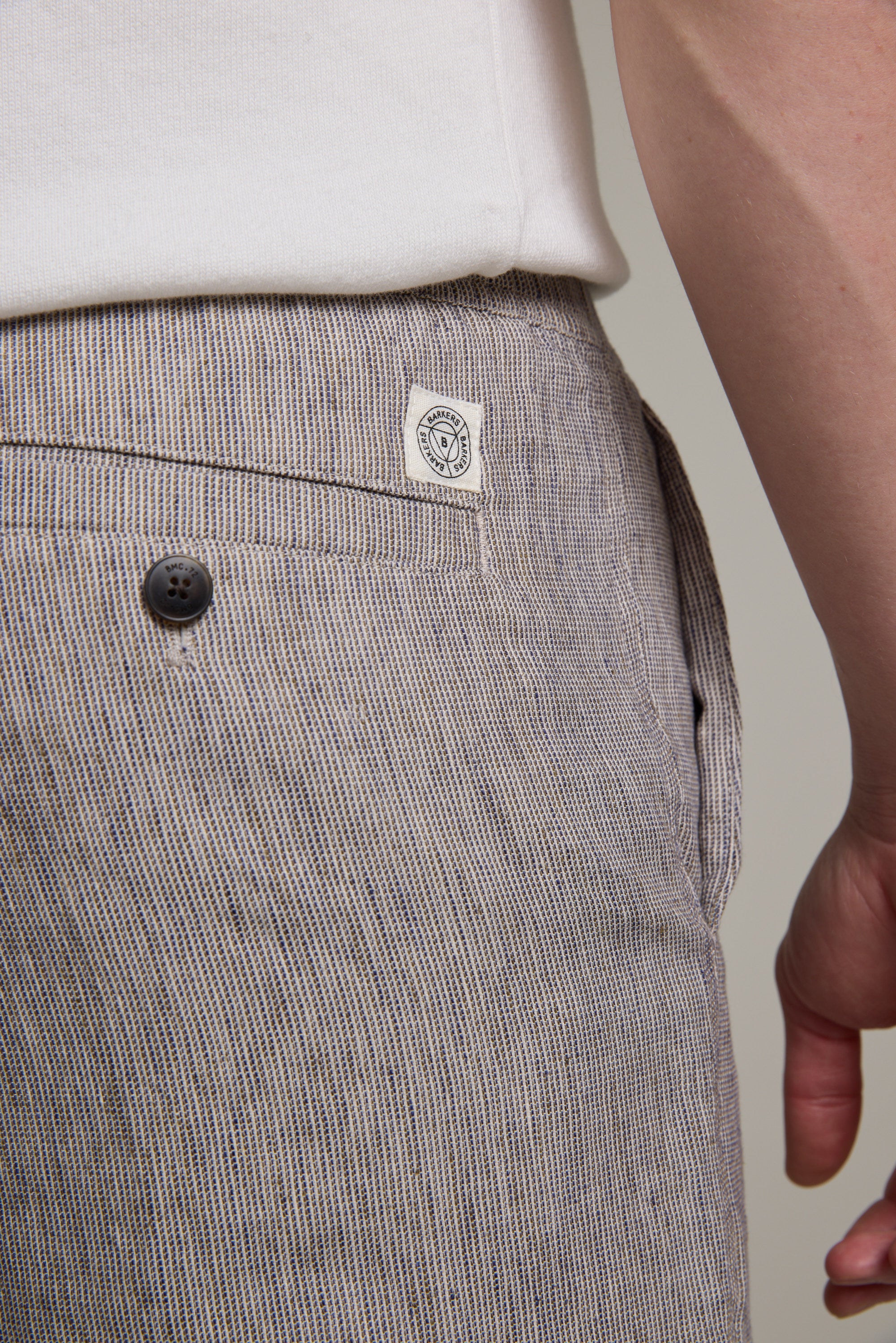 Close-up of a person wearing Barkers Woodley Weekend Shorts in light brown and white pinstripes, featuring a black buttoned back pocket, white stitched label, and visible part of a white shirt. The person's hand rests at their side.