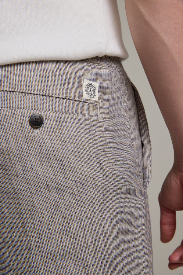 Close-up of a person wearing Barkers Woodley Weekend Shorts in light brown and white pinstripes, featuring a black buttoned back pocket, white stitched label, and visible part of a white shirt. The person's hand rests at their side.