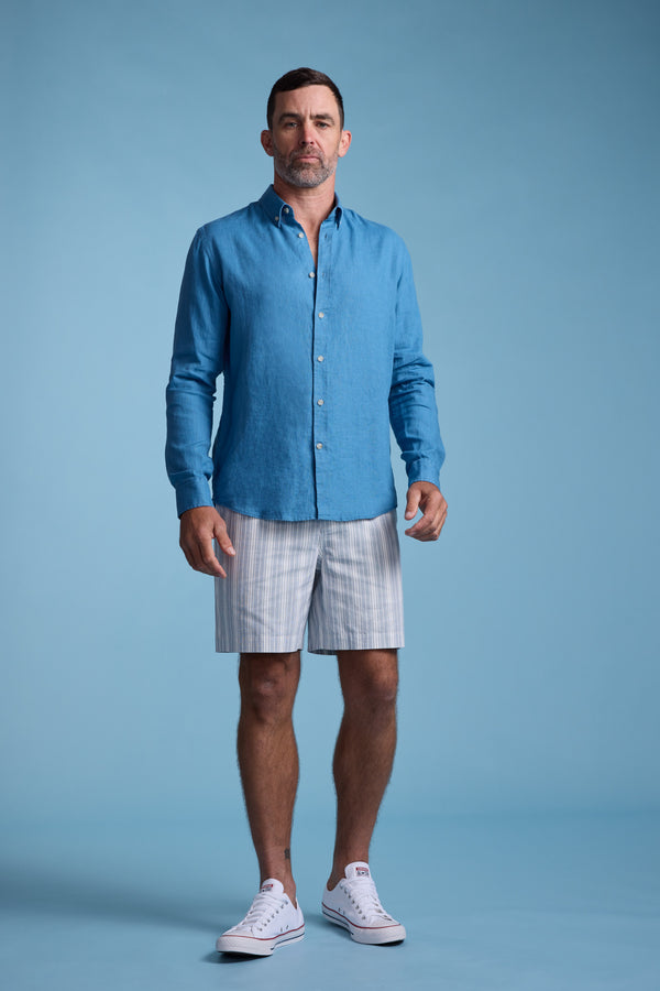 A man stands against a light blue background, wearing a blue button-up shirt, Barkers Manor Stripe Weekend Short with an elasticated waistband, and white sneakers. He faces forward with a neutral expression.