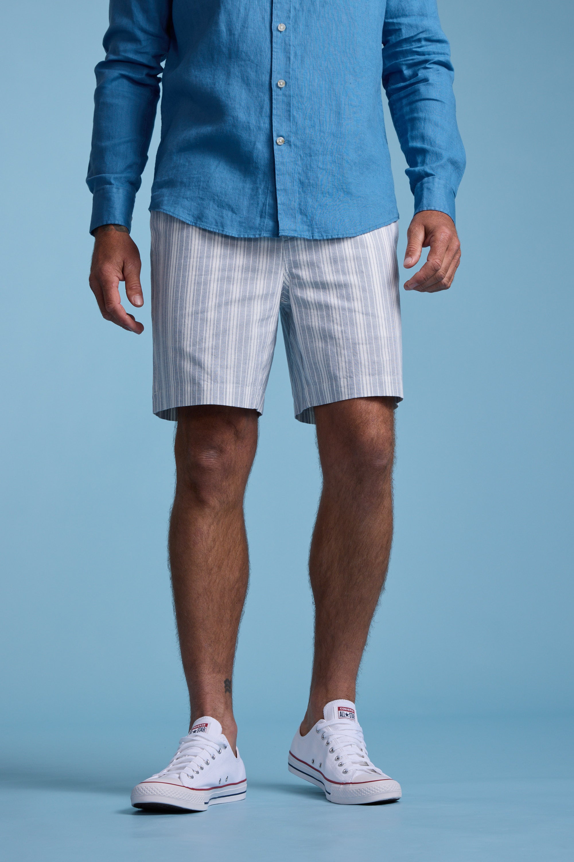 A man stands against a blue background, wearing a blue long-sleeve shirt, Barkers Manor Stripe Weekend Short with an elasticated waistband, and white sneakers. His face is not visible.
