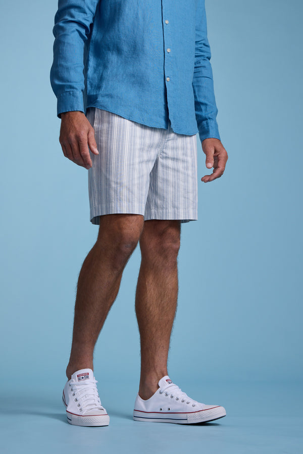 A man, visible from shoulders down, stands against a solid light blue background wearing Barkers Manor Stripe Weekend Shorts with an elasticated waistband, a light blue button-up shirt, and white sneakers.