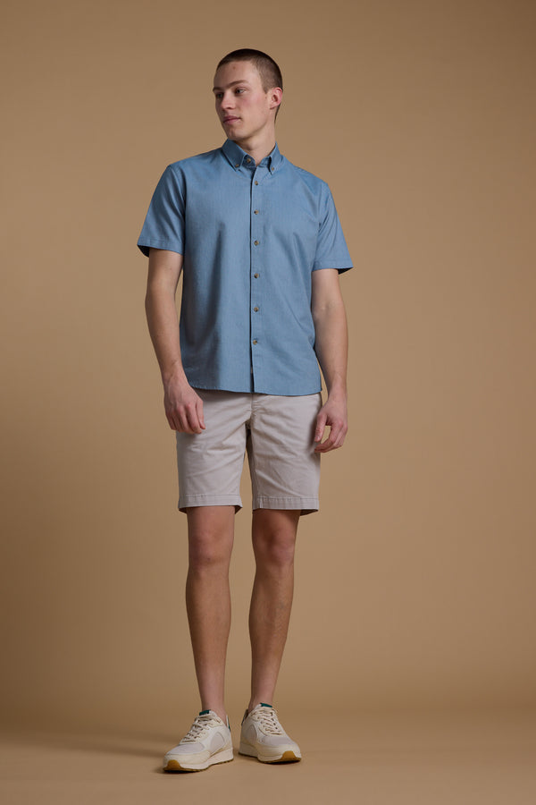 A person stands against a tan background wearing a light blue short-sleeved button-up, Barkers Kingly Chino Short men’s stretch shorts in light beige, and white sneakers, looking to the side with arms relaxed.