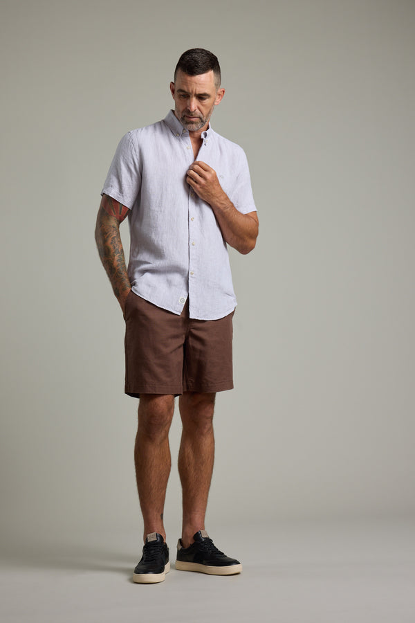 A man stands against a plain background, wearing the Barkers Gentry Linen Shirt in light grey, paired with brown shorts and black sneakers. He looks down, with one hand touching his shirt collar.