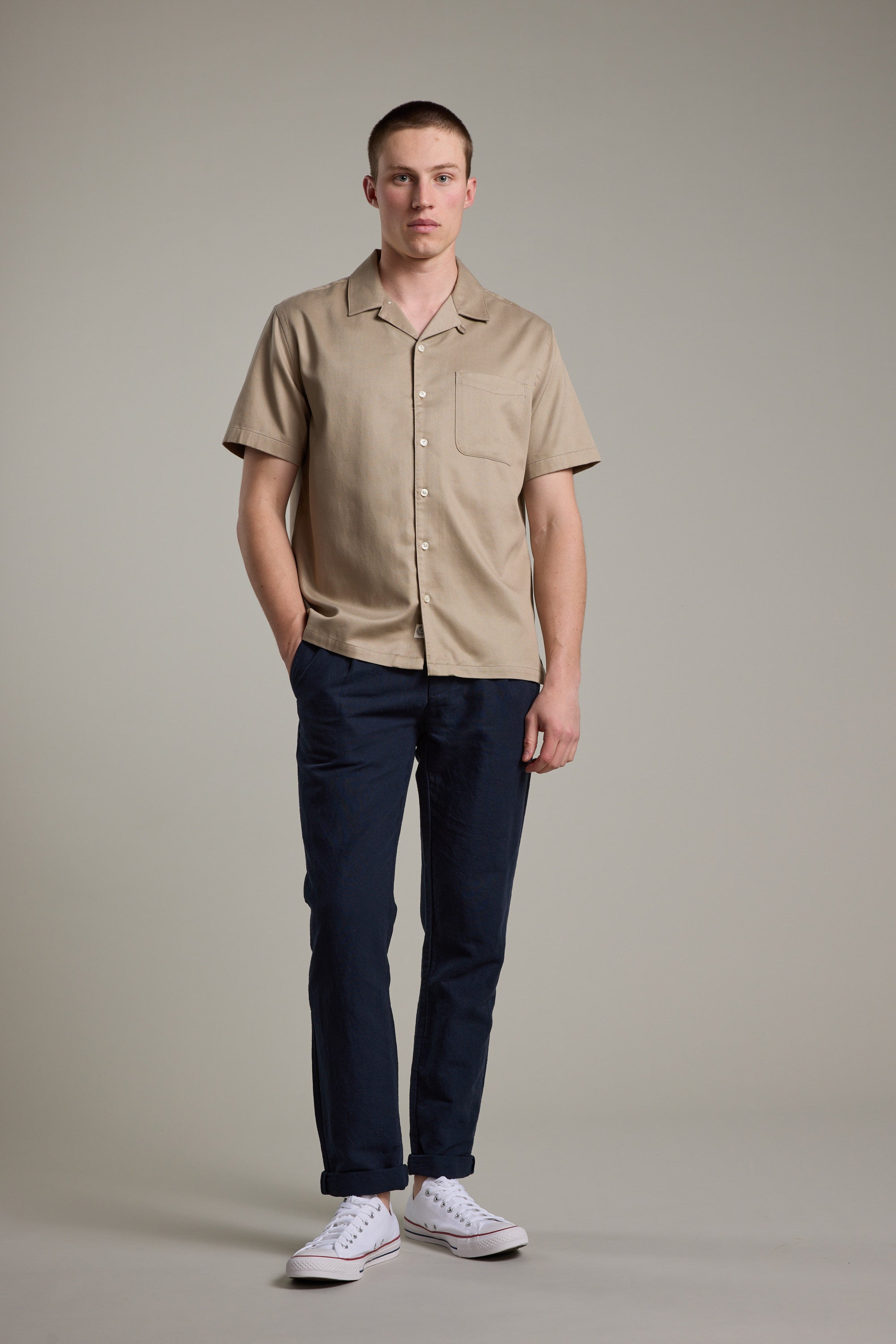 A man faces forward with a neutral expression, wearing the Barkers Augusta Cuban Shirt in tan—crafted from responsibly sourced cotton—paired with dark blue pants and white sneakers. One hand rests in his pocket against a plain background.
