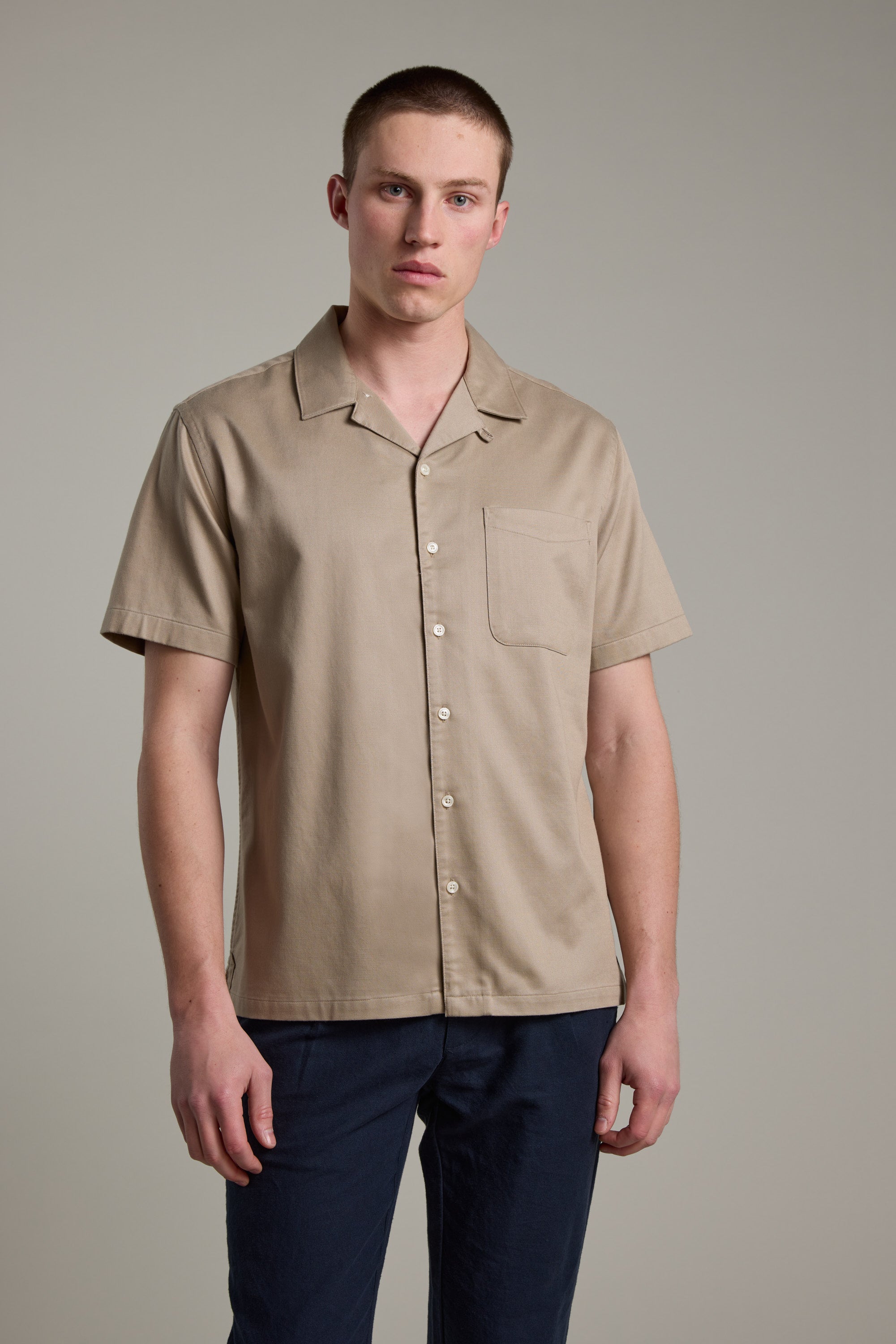 A young man with short hair wears the Barkers Augusta Cuban Shirt, a beige short-sleeve button-up with a chest pocket, made from responsibly sourced cotton. He stands against a plain light gray background in navy blue pants.
