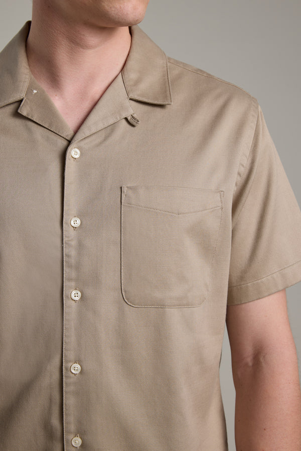 A person wears the Barkers Augusta Cuban Shirt in light brown, featuring short sleeves, a Cuban collar, and breast pocket. Shown from shoulders to mid-torso against a plain background. Made from responsibly sourced cotton.