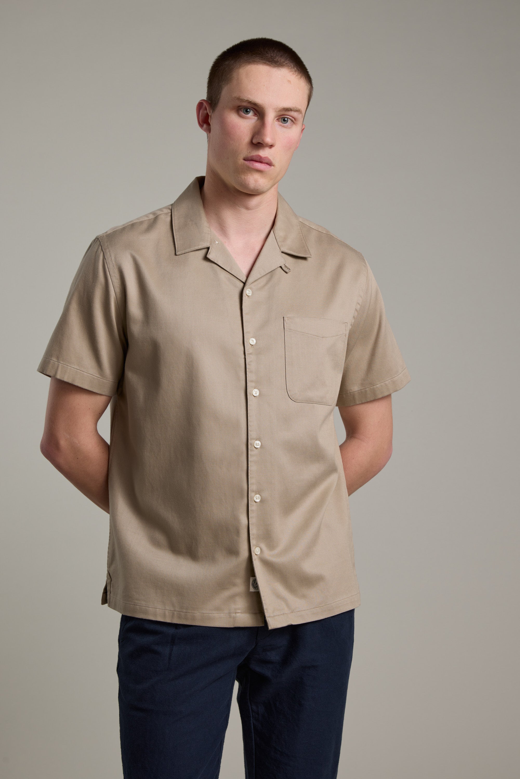 A young man with short hair wears the Barkers Augusta Cuban Shirt—made from responsibly sourced cotton, featuring a Cuban collar and chest pocket—with dark pants. He stands against a plain light gray background, hands behind his back.