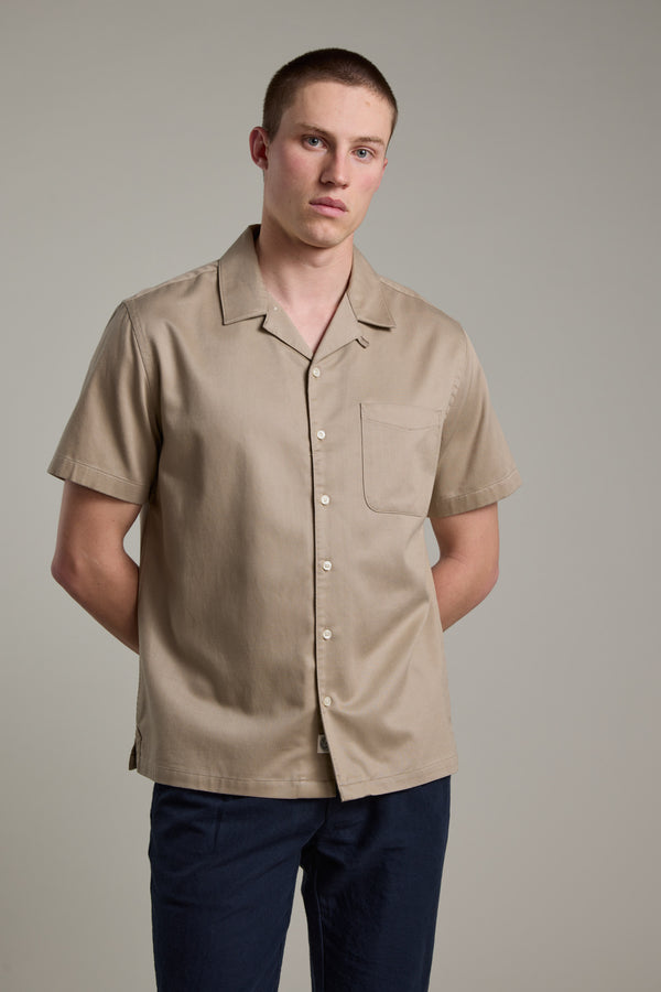 A young man with short hair wears the Barkers Augusta Cuban Shirt—made from responsibly sourced cotton, featuring a Cuban collar and chest pocket—with dark pants. He stands against a plain light gray background, hands behind his back.