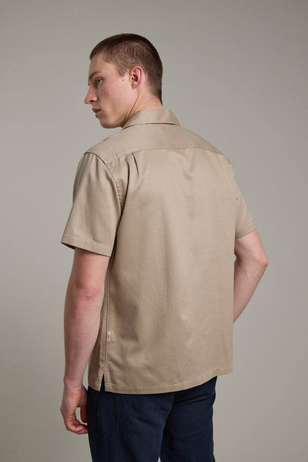 A man with short hair stands facing away, glancing over his shoulder in a beige Barkers Augusta Cuban Shirt made from responsibly sourced cotton and dark pants, set against a plain gray background.