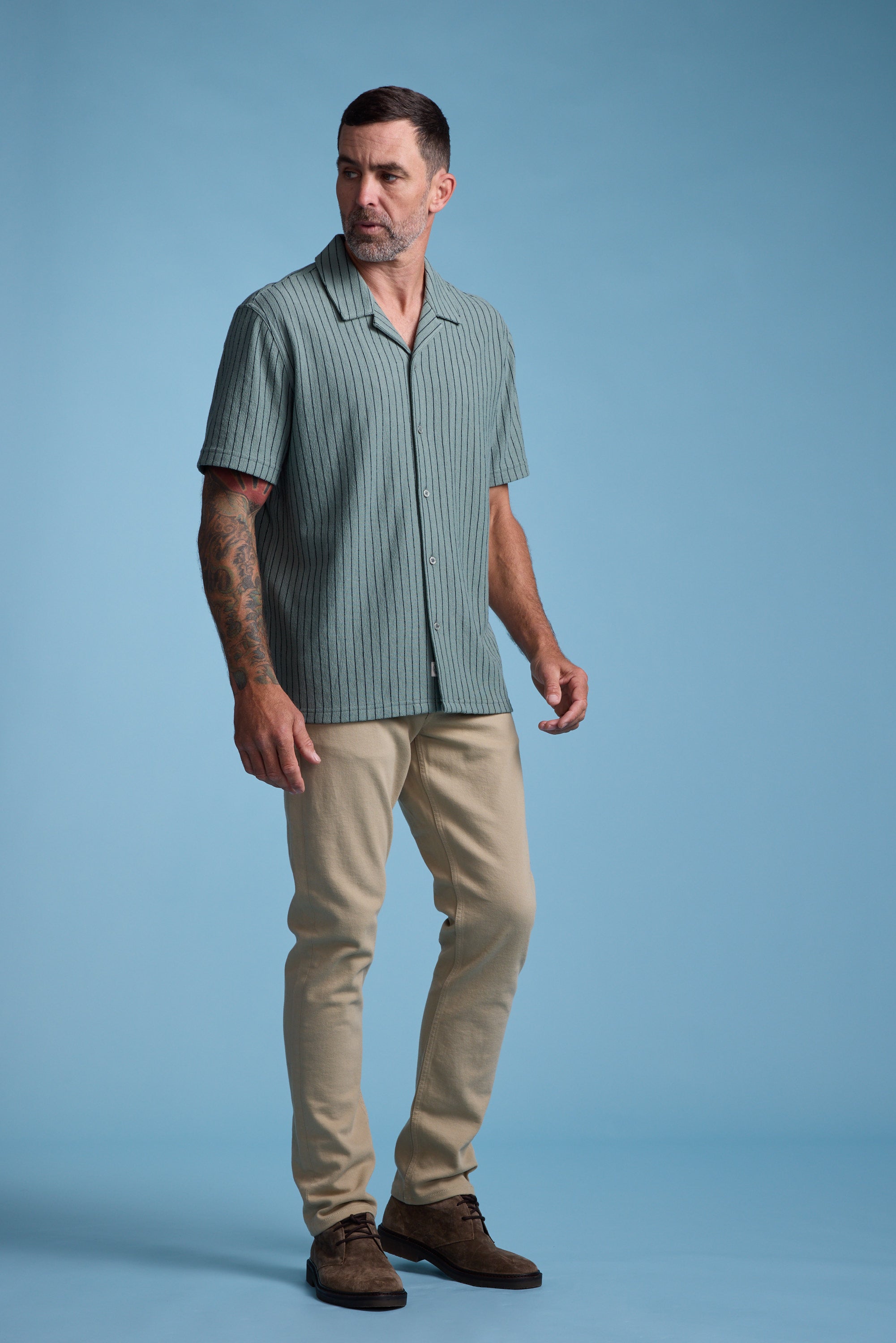 A man with short dark hair, a beard, and tattoos on his left arm stands against a light blue background, wearing the Barkers Diego Stripe Cuban Shirt in green terry toweling with short sleeves, paired with beige pants and brown shoes.