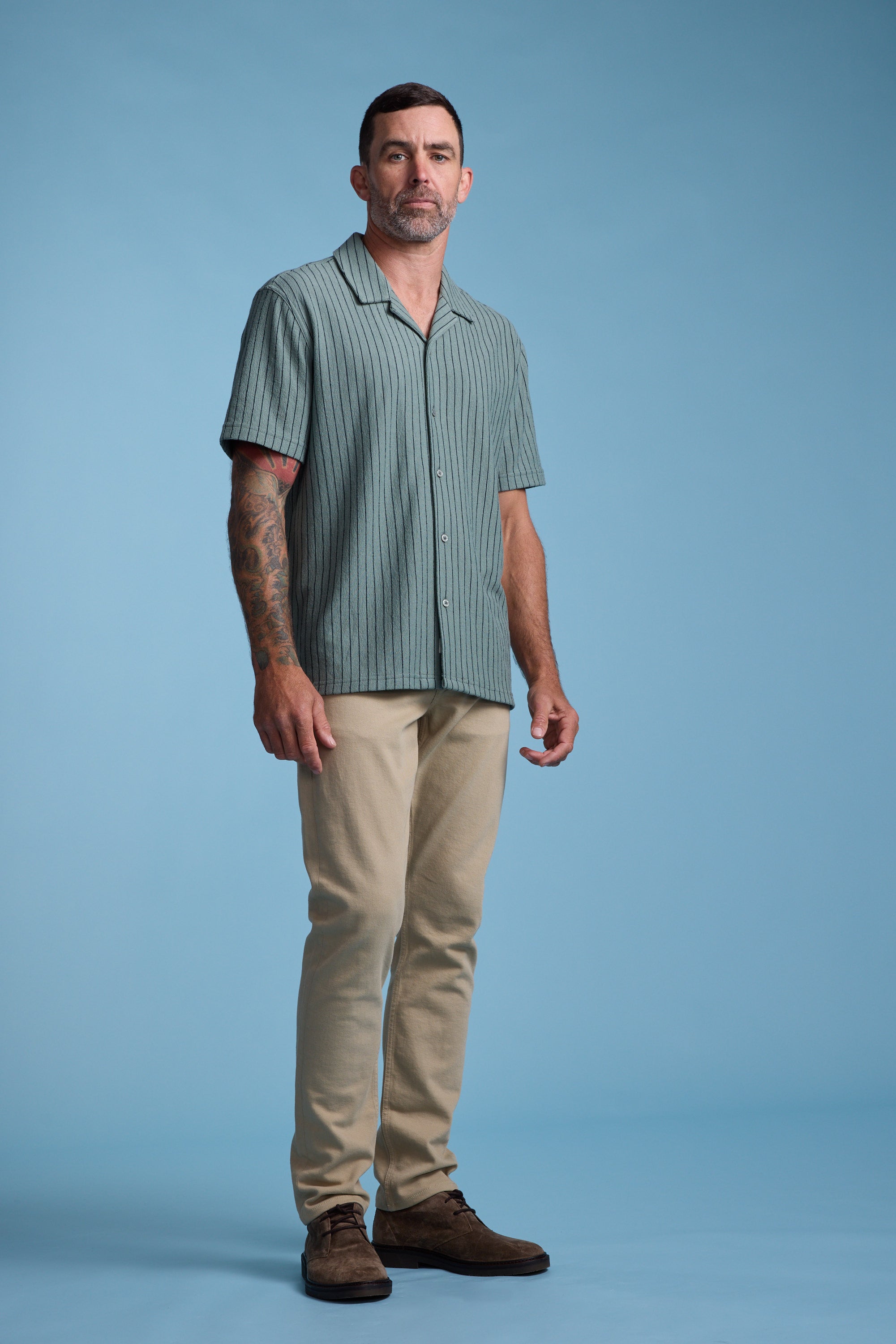 A man with short dark hair and tattoos on his left arm stands against a plain blue background, wearing the Barkers Diego Stripe Cuban Shirt, beige pants, and brown shoes.