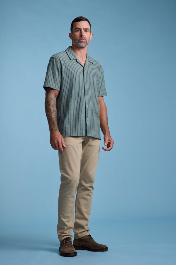 A man with short dark hair and tattoos on his left arm stands against a plain blue background, wearing the Barkers Diego Stripe Cuban Shirt, beige pants, and brown shoes.