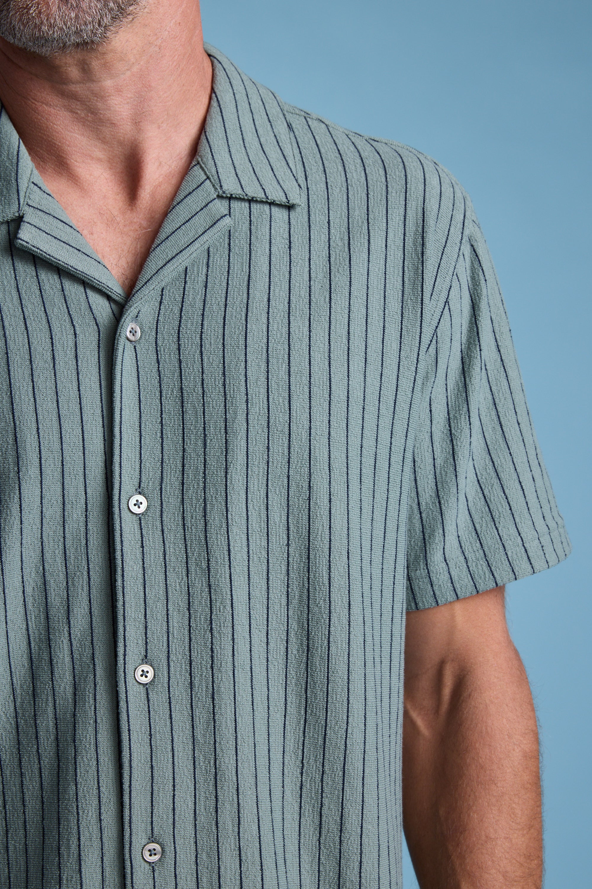 A close-up shows a man wearing the Barkers Diego Stripe Cuban Shirt, a light blue short-sleeved shirt with thin vertical stripes. Only his torso, left arm, and part of his face appear against a solid light blue background.