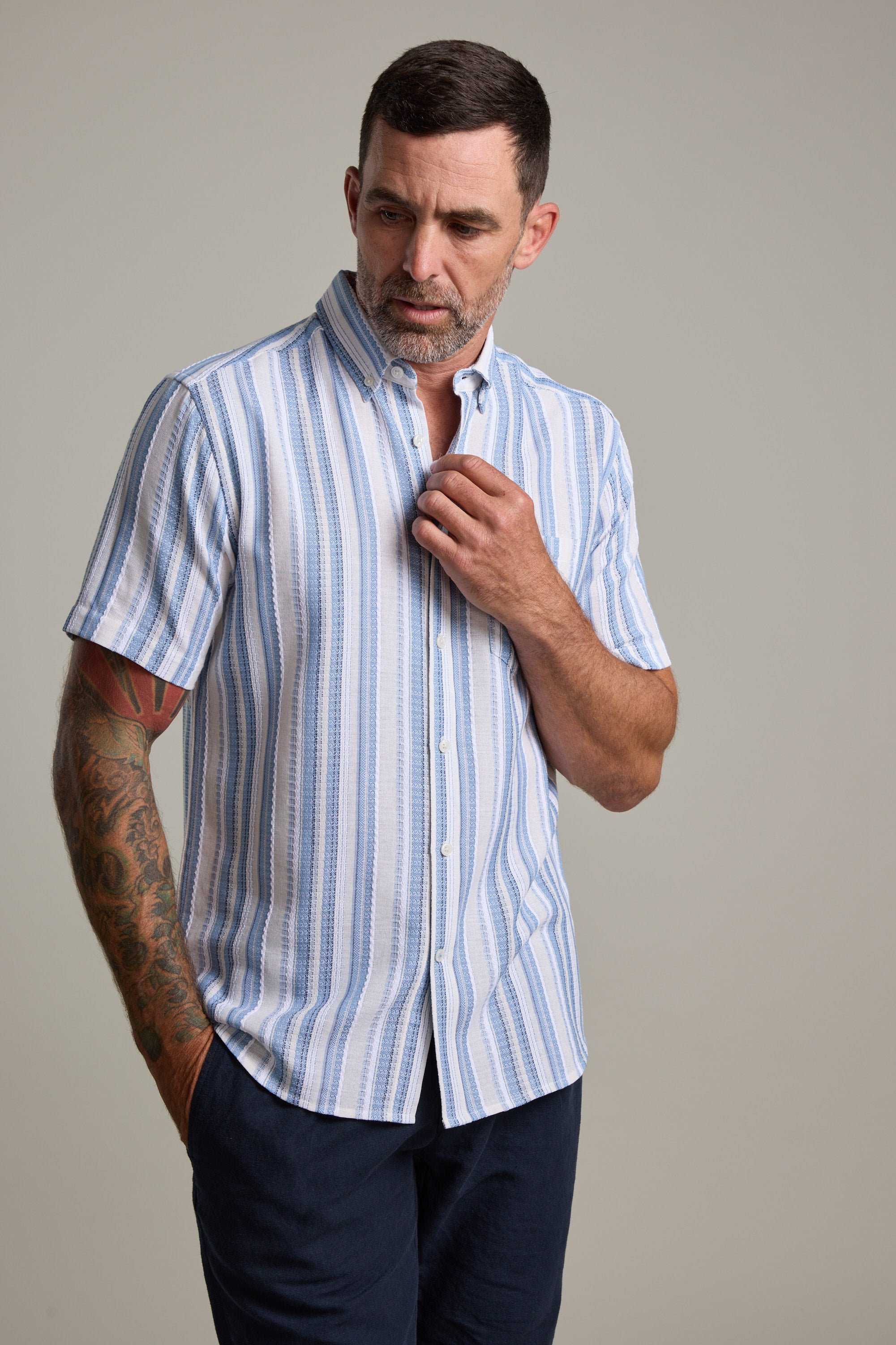 A man wears the Barkers Merten Stripe Shirt—short-sleeve, light blue and white cotton stripes—and dark pants, standing against a plain background. One hand is in his pocket as he adjusts his collar, showing a tattoo on his left forearm. Ideal for summer style.