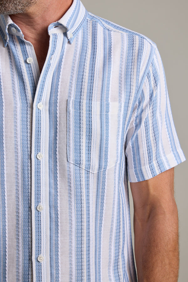 A man wears the Barkers Merten Stripe Shirt, a cotton short-sleeve with blue and white vertical stripes and a chest pocket. The photo shows his upper torso against a plain background, making it ideal for any summer wardrobe.