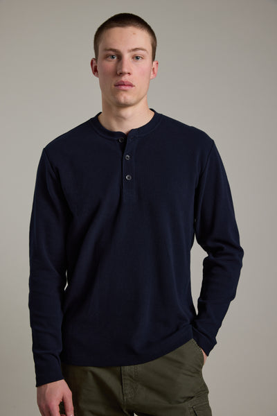 A young man with short hair wears the Barkers Avenue Waffle Henley in navy blue, made from organic cotton, and olive green pants, standing against a plain light gray background.