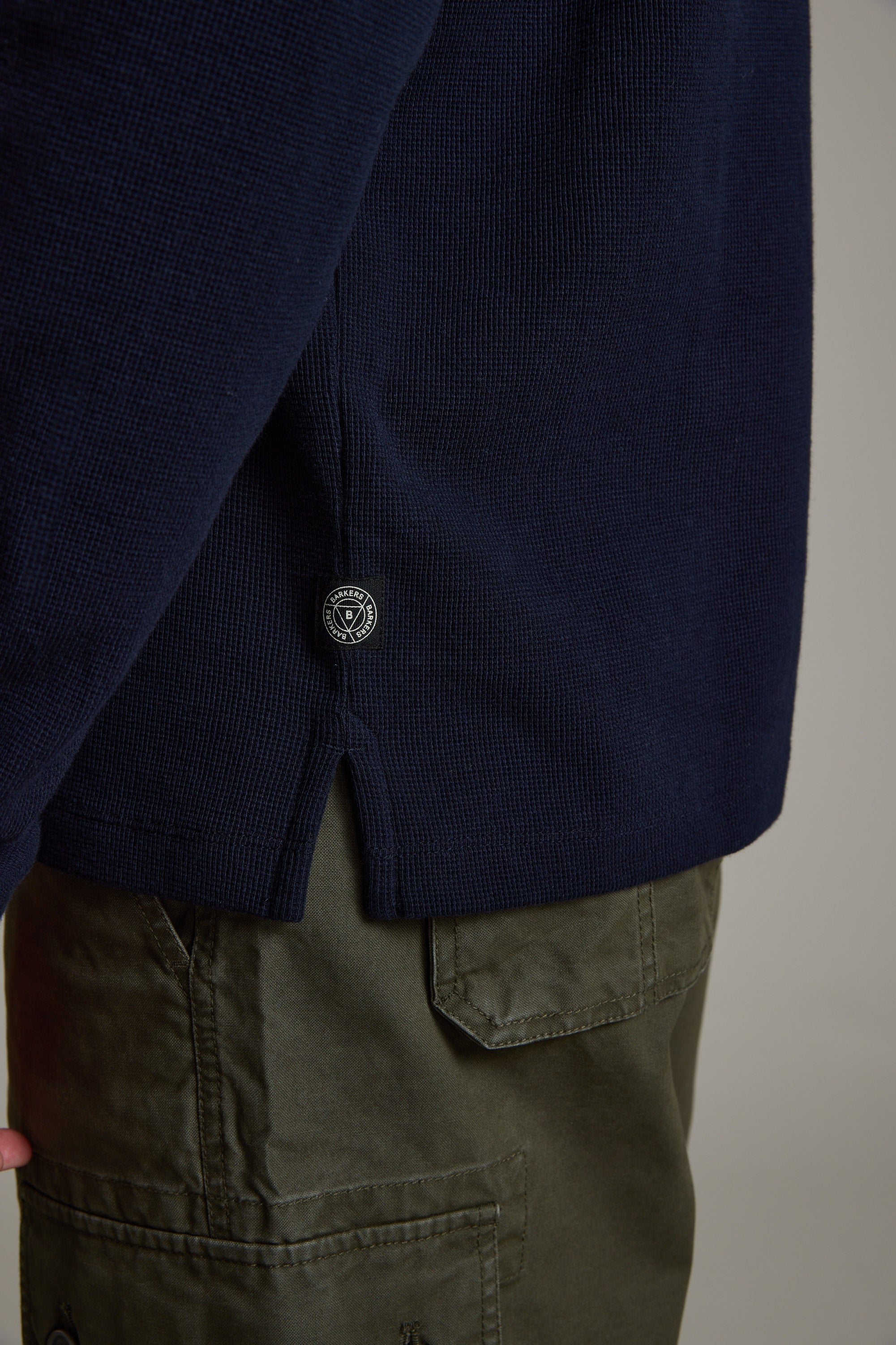 A close-up of a person wearing olive green pocketed pants and the Avenue Waffle Henley by Barkers, a men's mid-weight navy blue organic cotton sweater featuring a small round black and white tag near the hem.