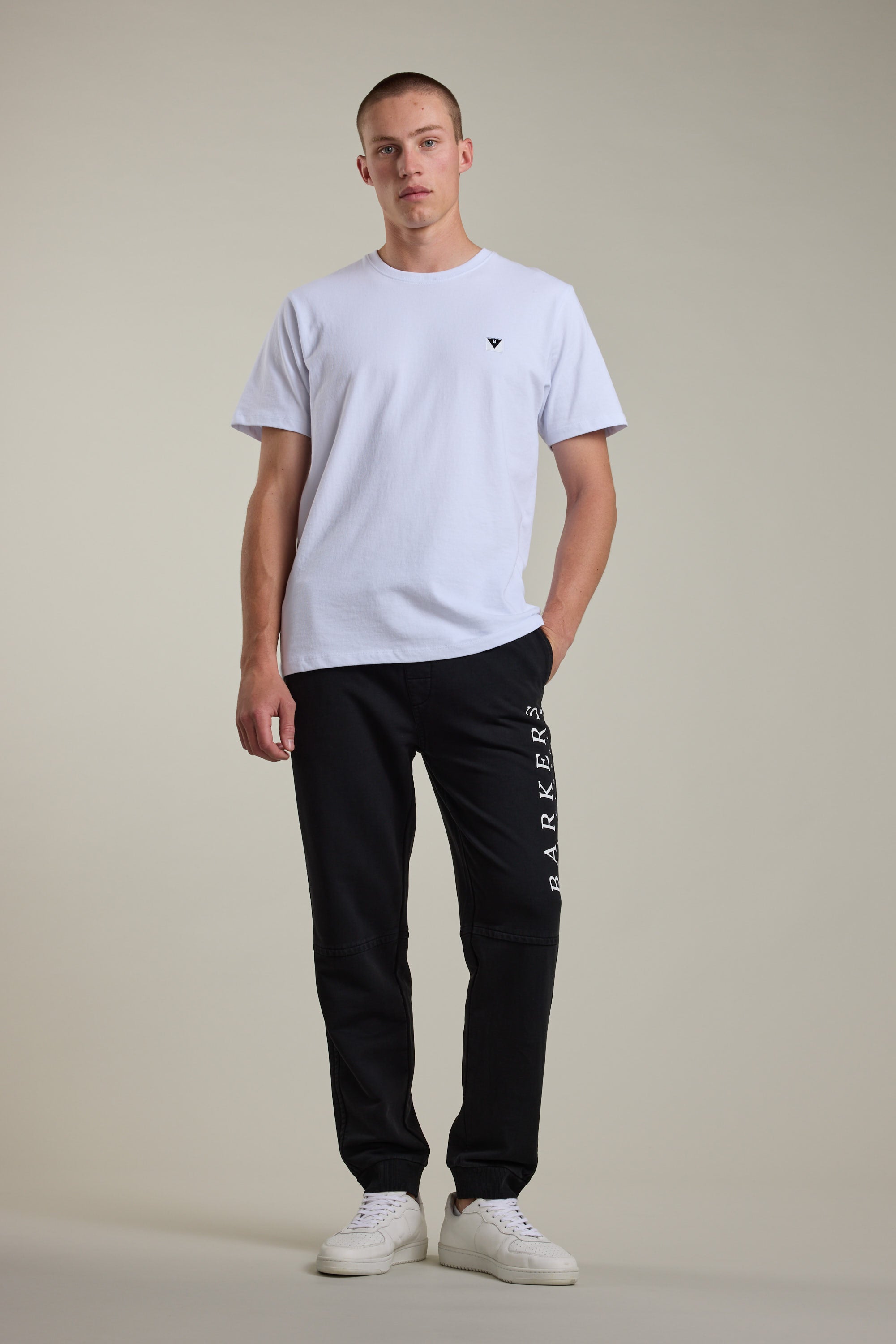 A man stands facing forward against a plain background, wearing a white short-sleeve t-shirt, Barkers Organic Classic Print Track Pant with leg text, and white sneakers; one hand is in his pocket.