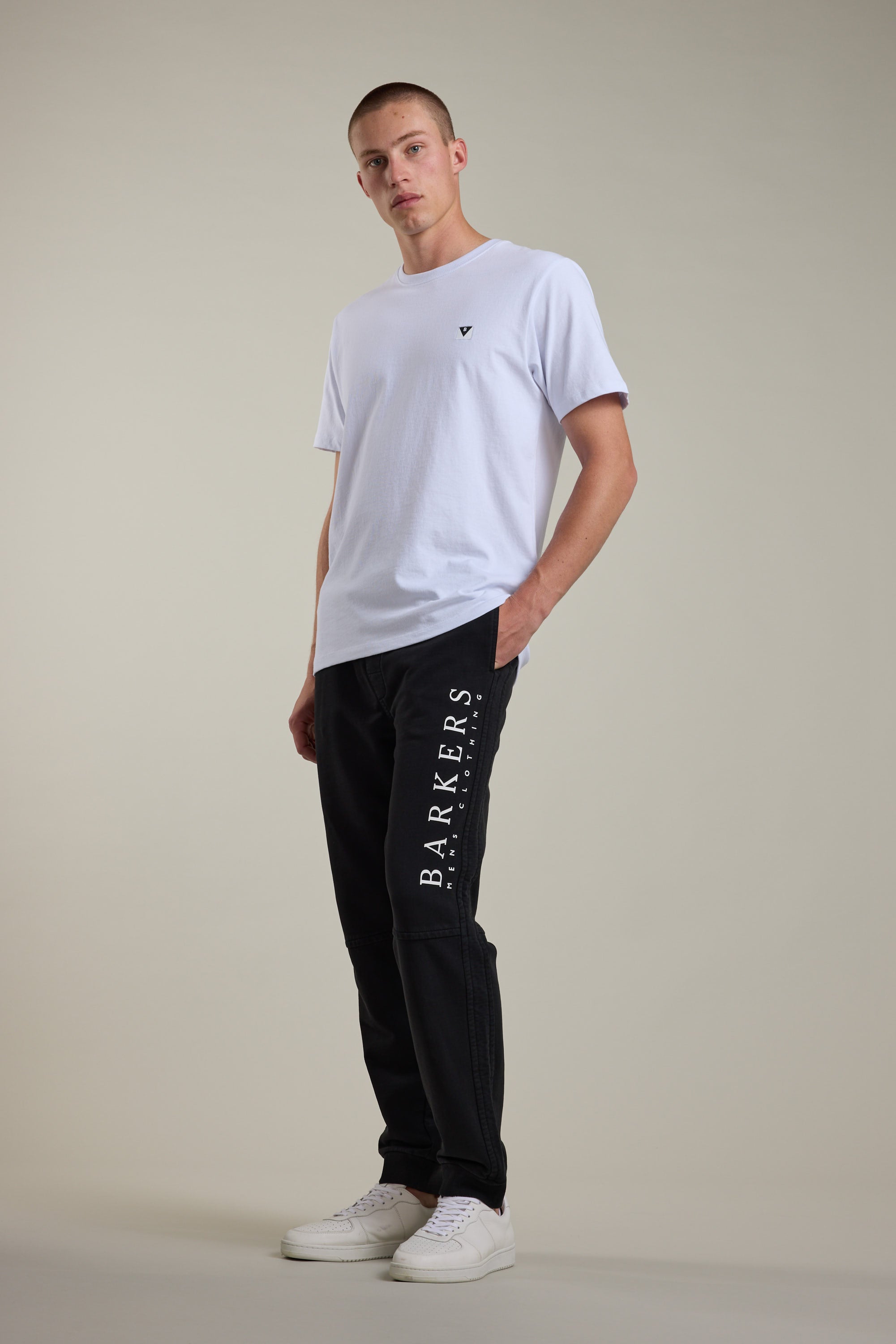 A man stands in a relaxed pose wearing a plain white T-shirt, Barkers Organic Classic Print Track Pant with "BARKERS" printed down the leg in white, and white sneakers against a neutral background.