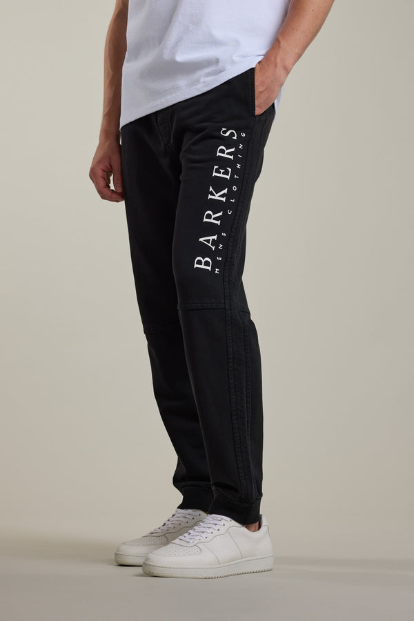 A person wears black Barkers Organic Classic Print Track Pants with "BARKERS MENS CLOTHING" in white on the left thigh, a white tee, and white sneakers against a plain background—classic ‘90s-inspired style with a modern twist.