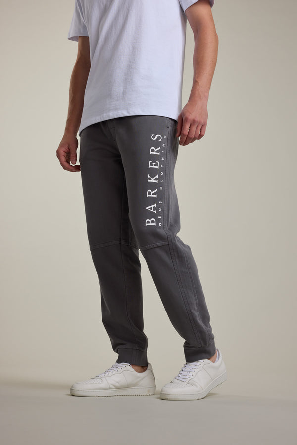 A person wears a white t-shirt, Barkers Organic Classic Print Track Pant in grey with BARKERS text on the leg, and white sneakers, standing against a plain light background.