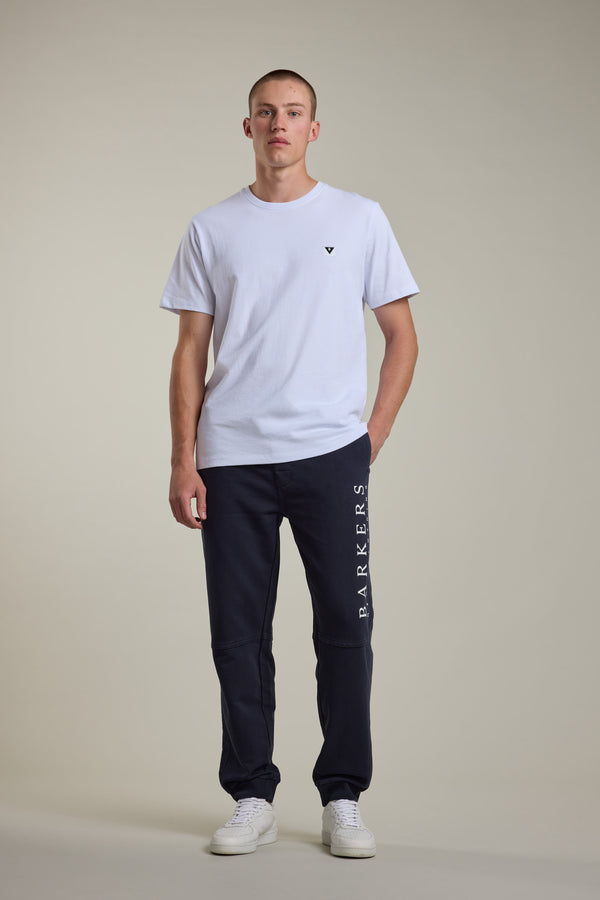 A man stands against a plain background wearing a white t-shirt, Barkers Organic Classic Print Track Pant with a logo on the leg, and white sneakers. One hand is in his pocket, and he has a neutral expression.