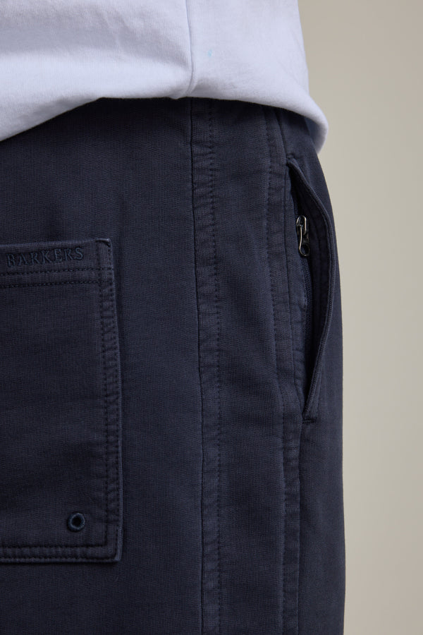 Close-up of a person wearing Barkers Organic Classic Print Track Pant in navy, featuring a back pocket, visible stitching, and the Barkers brand label above the pocket—evoking a classic '90s trackies vibe. Paired with a white shirt.