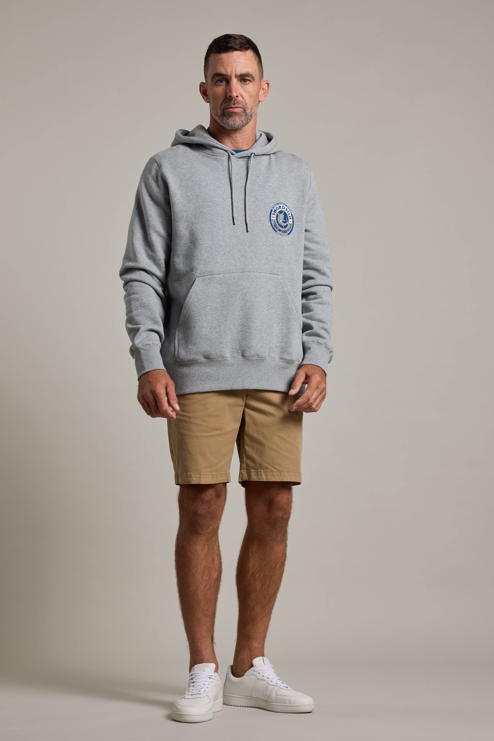 A man stands facing forward against a plain light background, wearing a light gray Swordfish Mojo Logo Hoody with a front pocket and chest logo, tan shorts, and white sneakers.