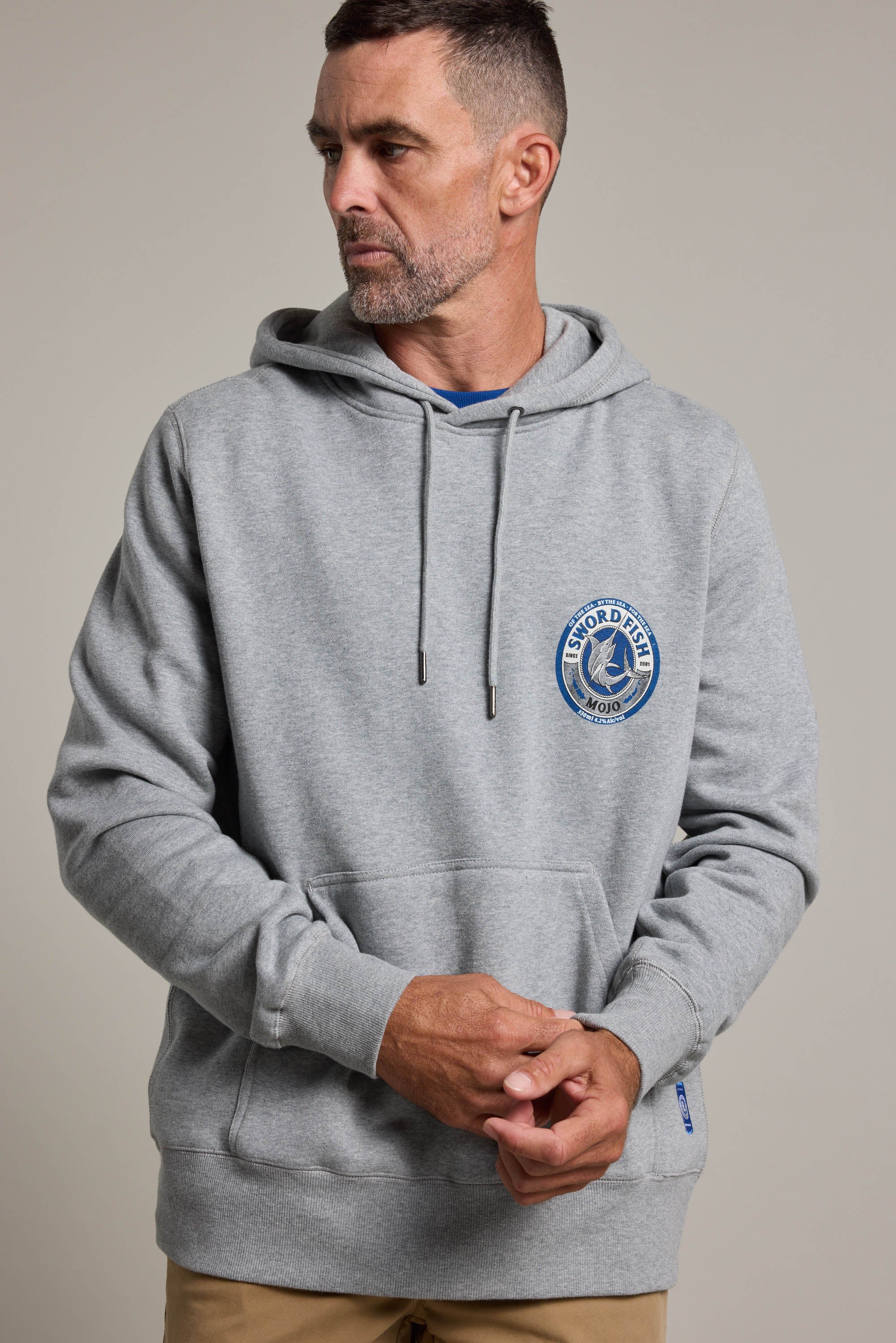 A man with short hair and a beard wears the light gray Swordfish Mojo Logo Hoody by Swordfish Mojo, featuring a circular Oxford Point logo on the chest. He stands with hands clasped, looking to the side against a plain background.