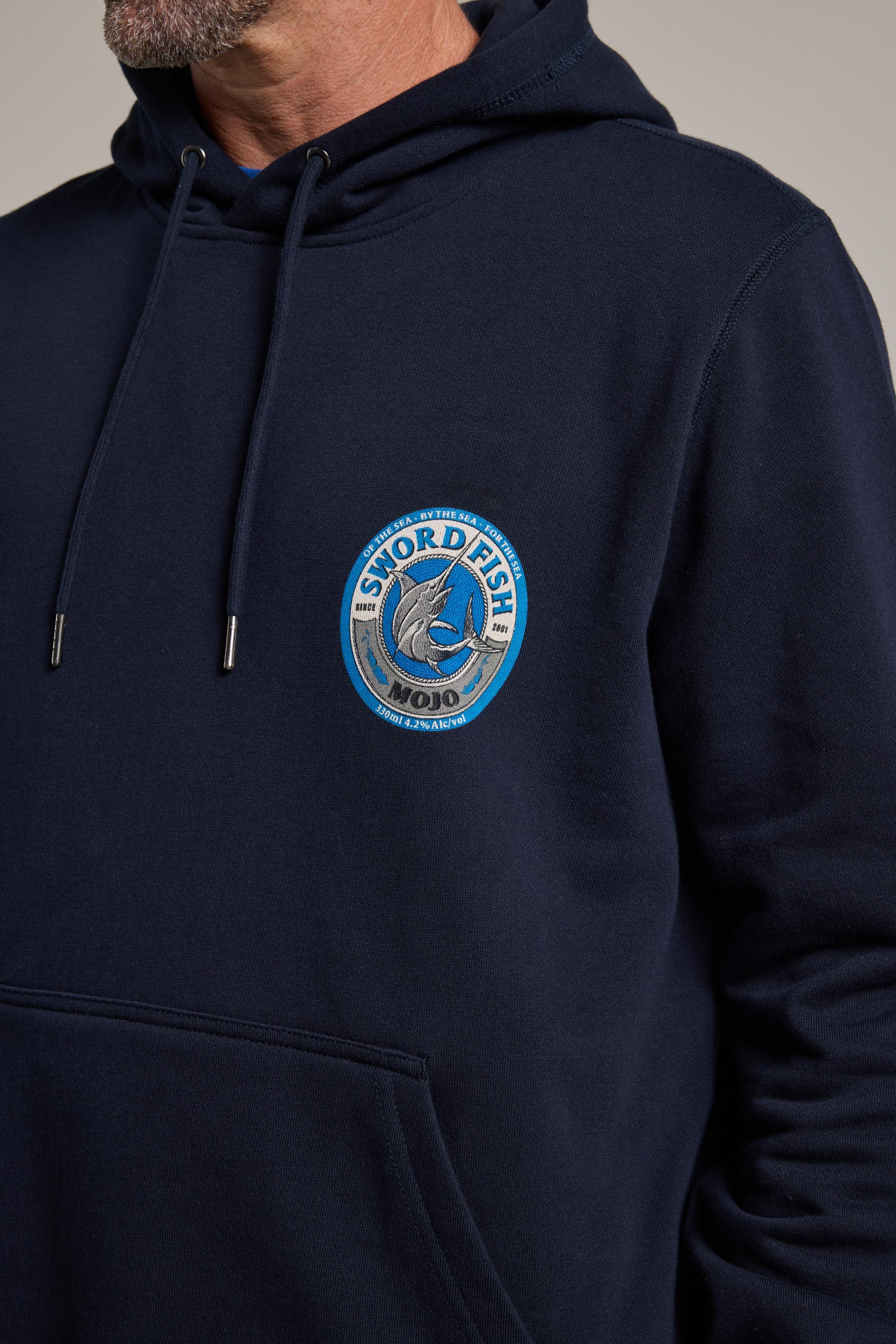 A man wears a Swordfish Mojo Logo Hoody by Swordfish Mojo, made from responsibly sourced cotton, with a chest patch featuring a blue and white swordfish logo. Only the lower half of his face is visible.
