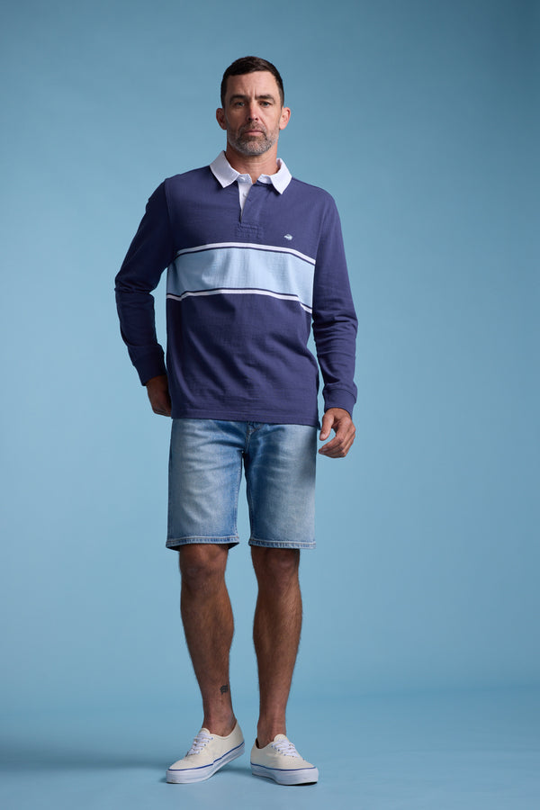 A man stands against a blue backdrop wearing the Barkers Breaker Rugby Jersey in navy with white and light blue stripes, paired with light blue denim shorts and white slip-on sneakers.