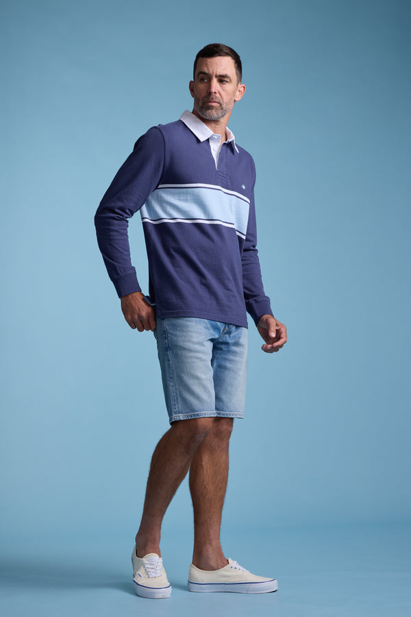 A man stands against a blue background wearing the Barkers Breaker Rugby Jersey in navy and light blue stripes, paired with light denim shorts and white slip-on shoes. He looks to his left with a neutral expression.