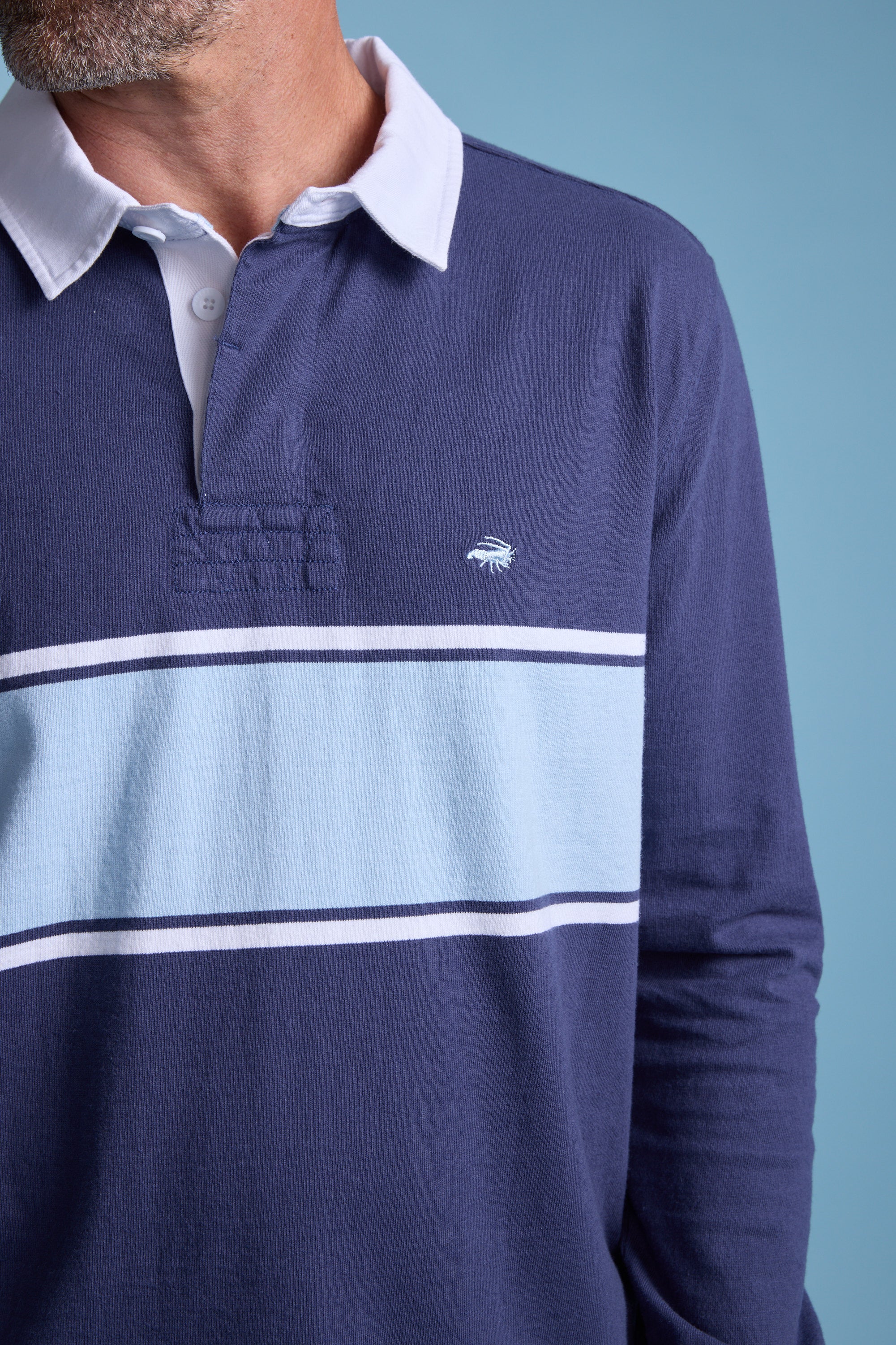 A man wears the Barkers Breaker Rugby Jersey, navy with a white collar, light blue chest stripe bordered by thin white lines, and a small embroidered logo. Made from organic cotton. Light blue background.