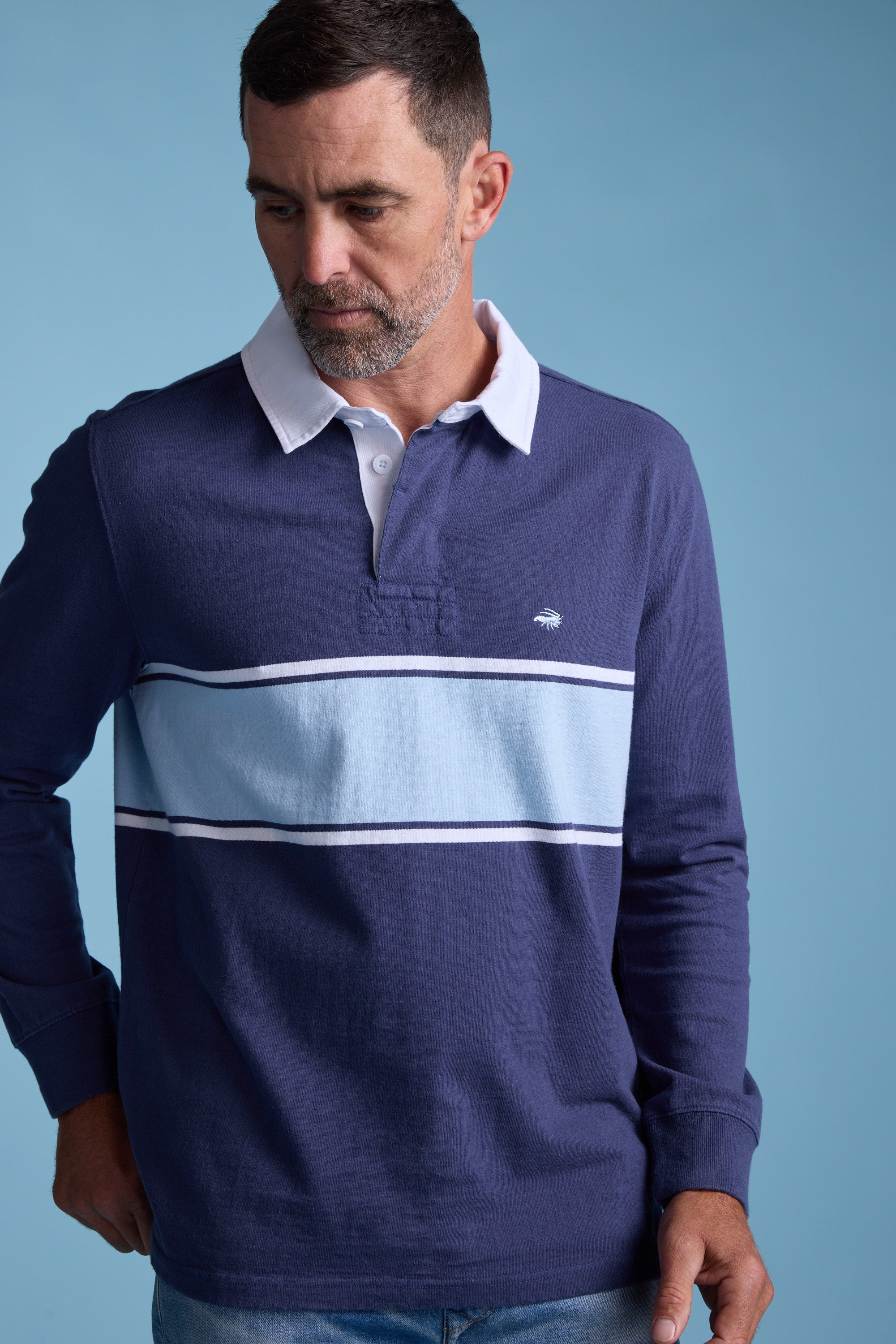 A man with short dark hair and a beard wears the Barkers Breaker Rugby Jersey—navy blue and white with horizontal stripes—crafted from organic cotton, standing against a light blue background and looking down to his left.