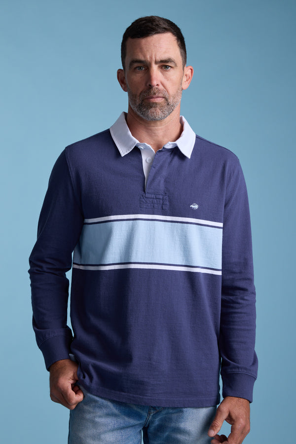A man with short dark hair and a trimmed beard wears the Barkers Breaker Rugby Jersey—navy blue with light blue and white stripes, a white collar, and made from organic cotton—standing against a plain blue background.